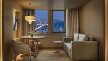 A modern hotel room with a dining area, a sofa, and a view of a cityscape with a bridge.