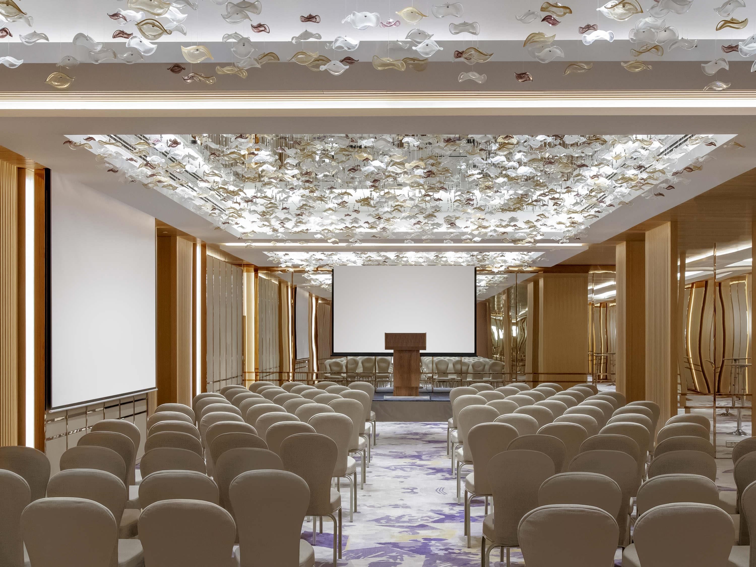 A conference room with rows of chairs, a podium, and a ceiling with hanging light fixtures.