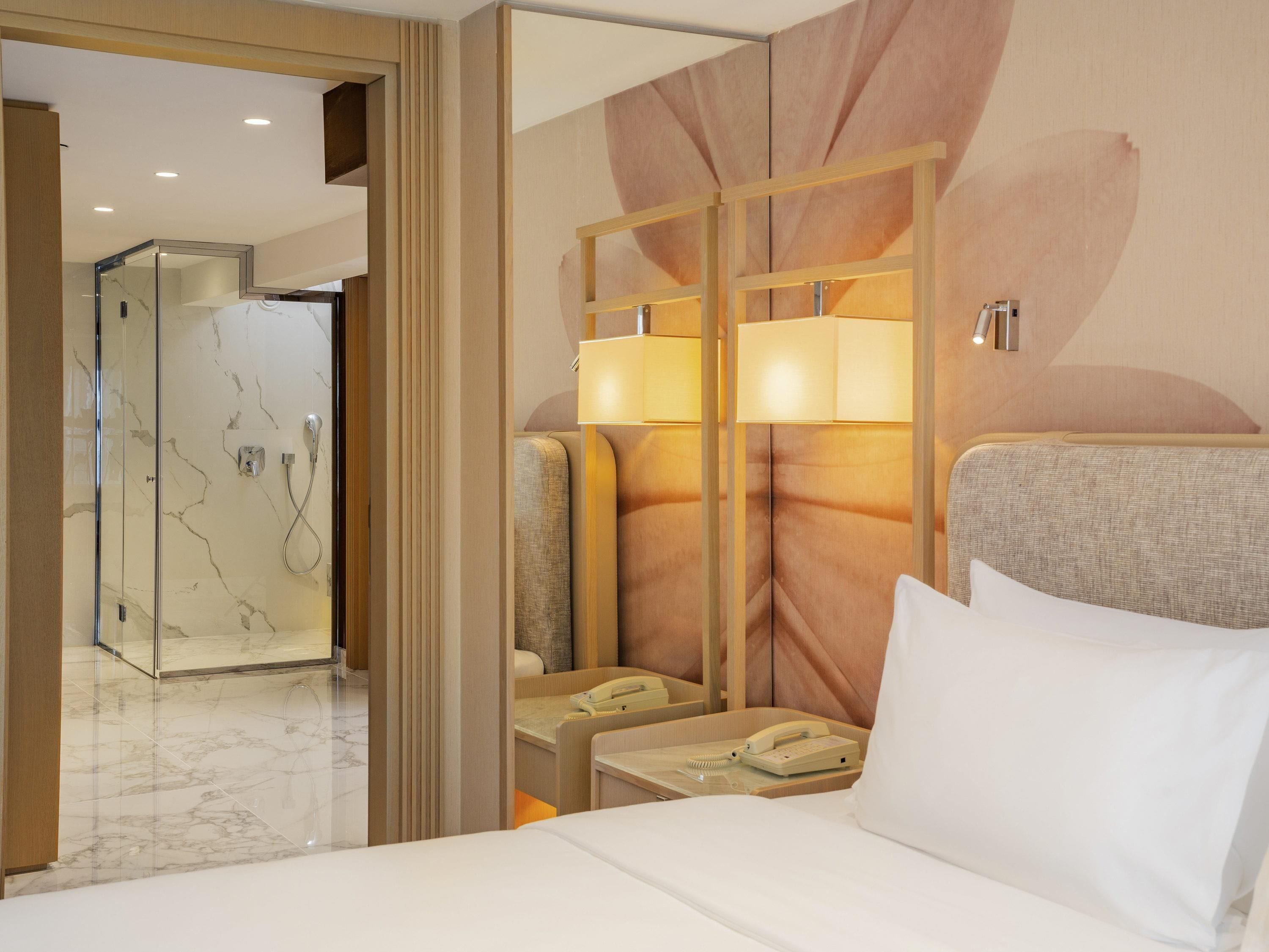 A hotel room with a bed, a bedside table with a lamp, a telephone, and a shower area with glass doors.