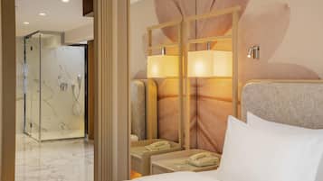 A hotel room with a bed, a bedside table with a lamp, a telephone, and a shower area with glass doors.