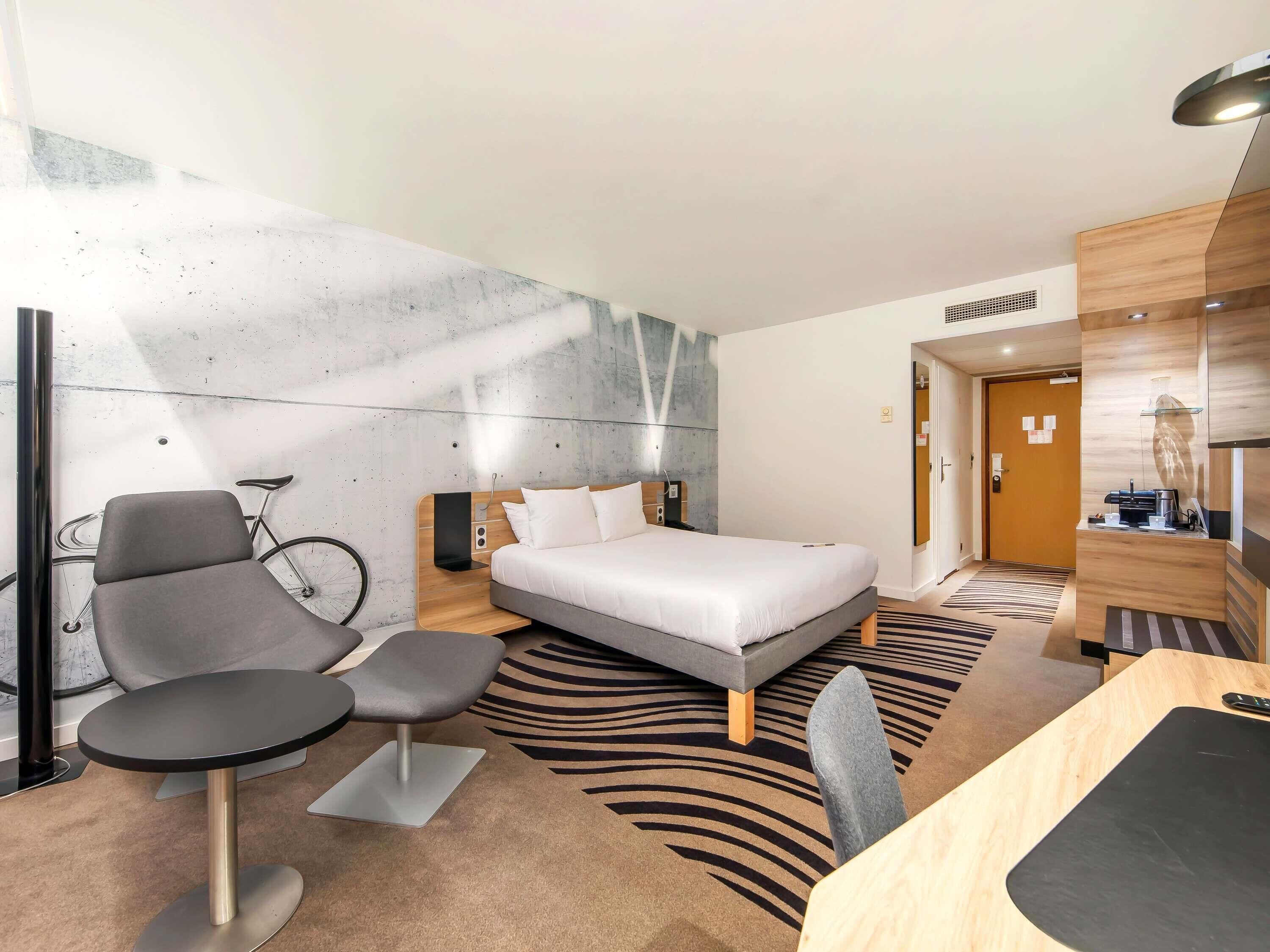 Una habitación de hotel moderna con cama, escritorio, silla, bicicleta y mostrador de recepción.