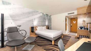 A modern hotel room with a bed, a desk, a chair, a bicycle, and a reception desk.