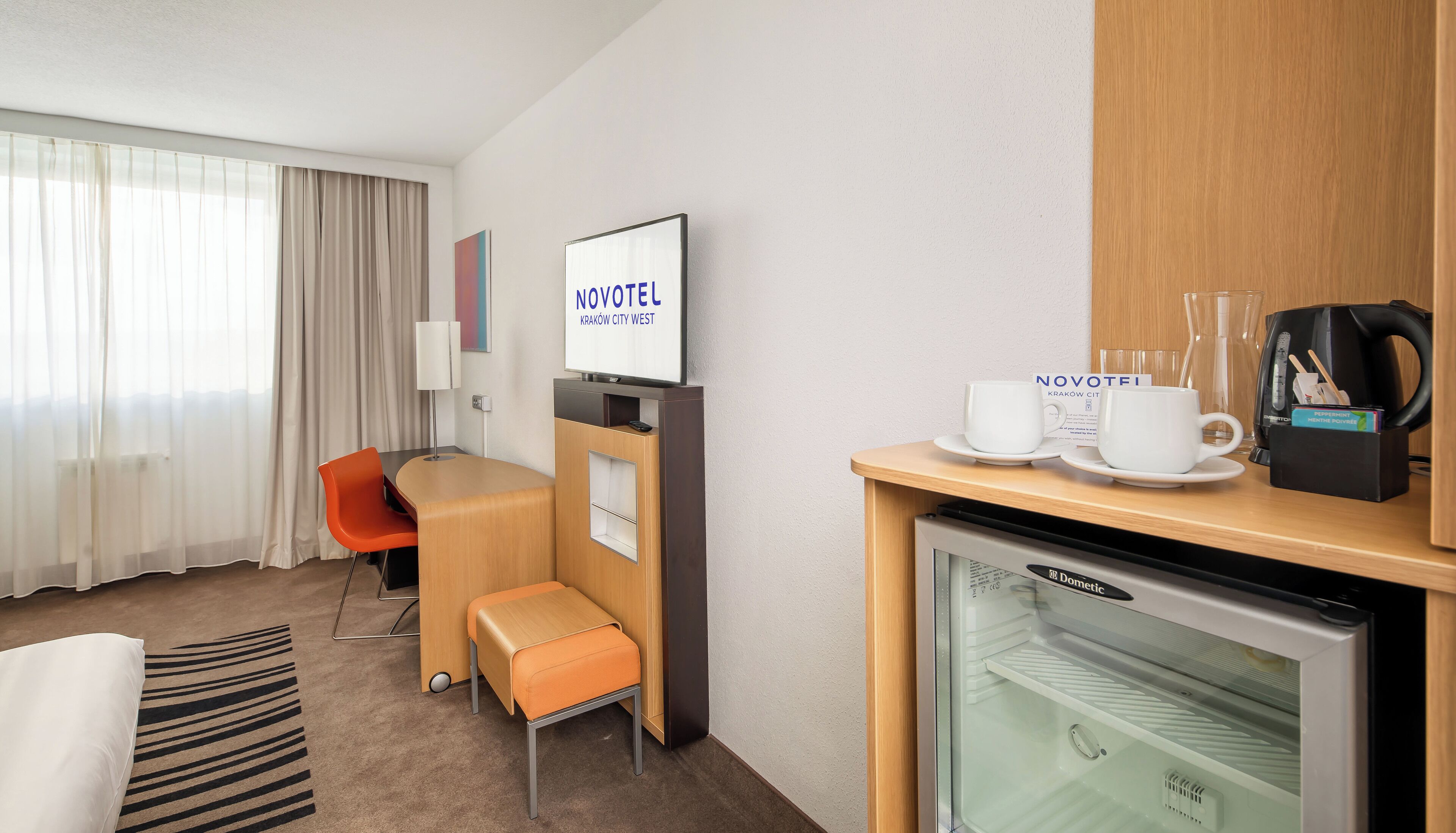 A hotel room with a bed, a desk with a chair, a coffee machine, and a TV displaying 'NOVOTEL'.