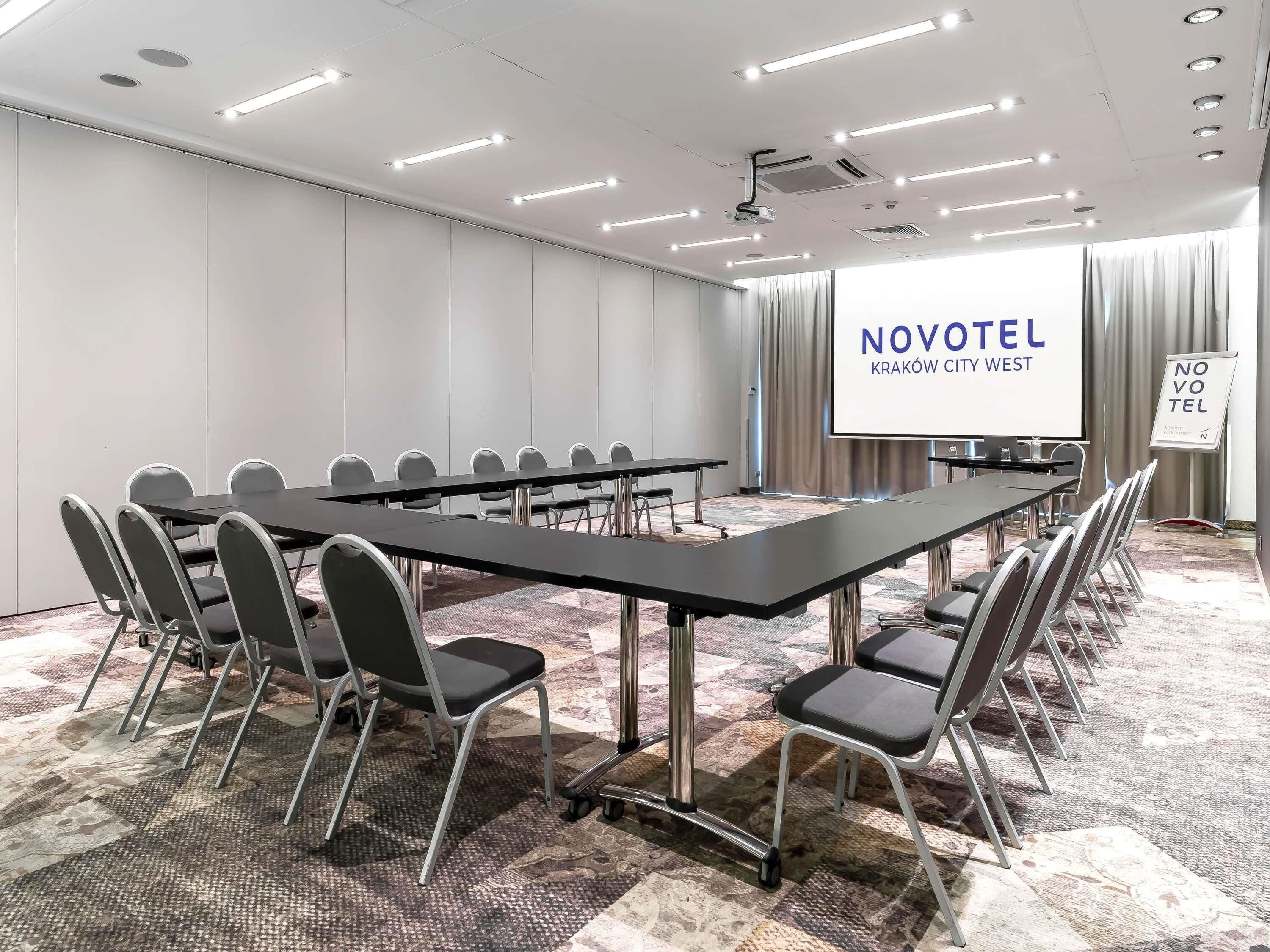 A conference room with a long table, chairs, a projector screen, and a sign for Novotel Kraków City West.
