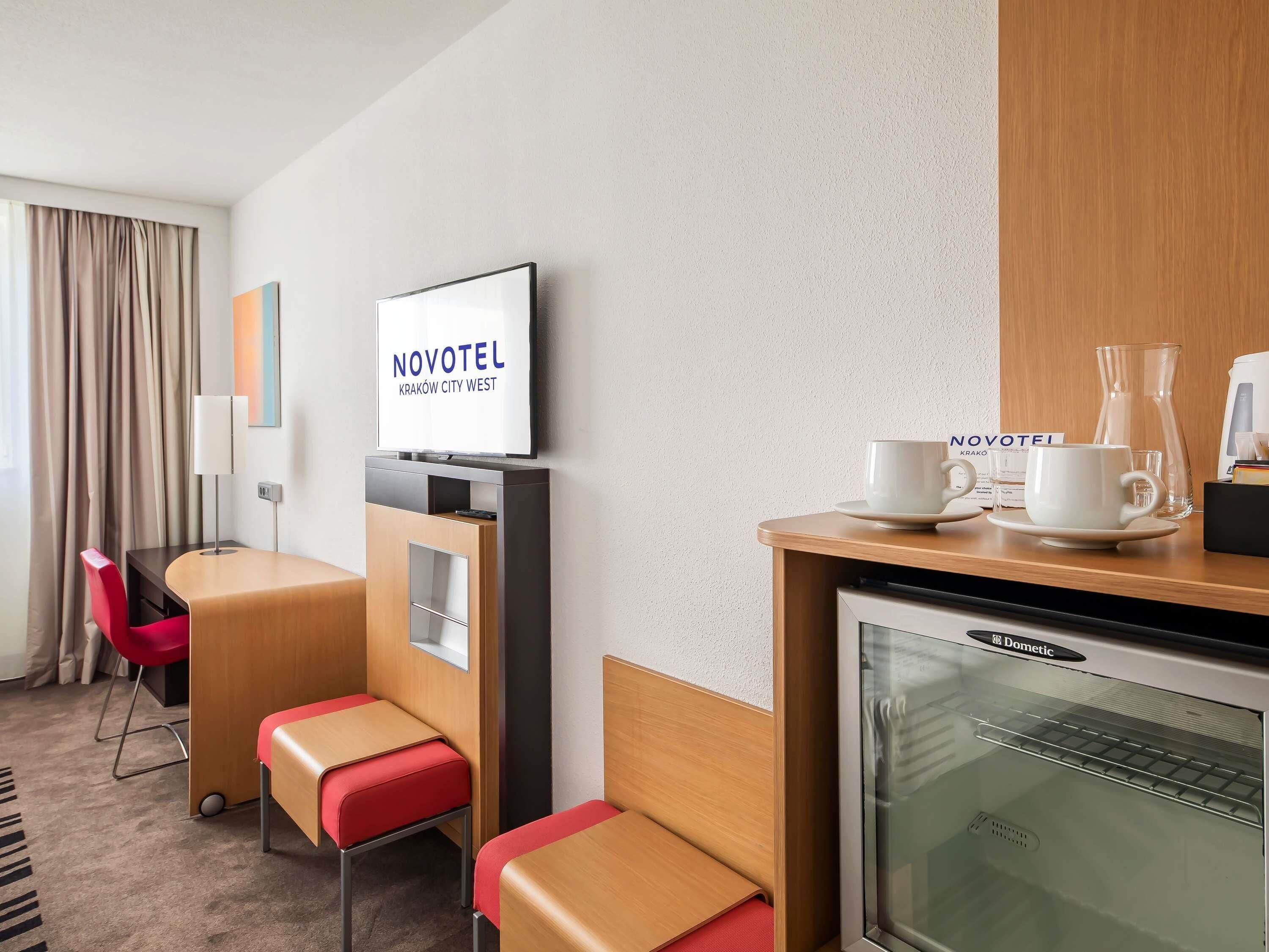 Habitación de hotel con televisión, minibar, estación de café y una zona de estar con sillón y reposapiés rojos.