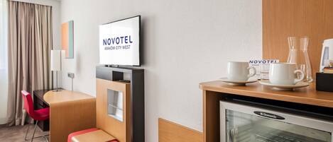 Habitación de hotel con televisión, minibar, estación de café y una zona de estar con sillón y reposapiés rojos.