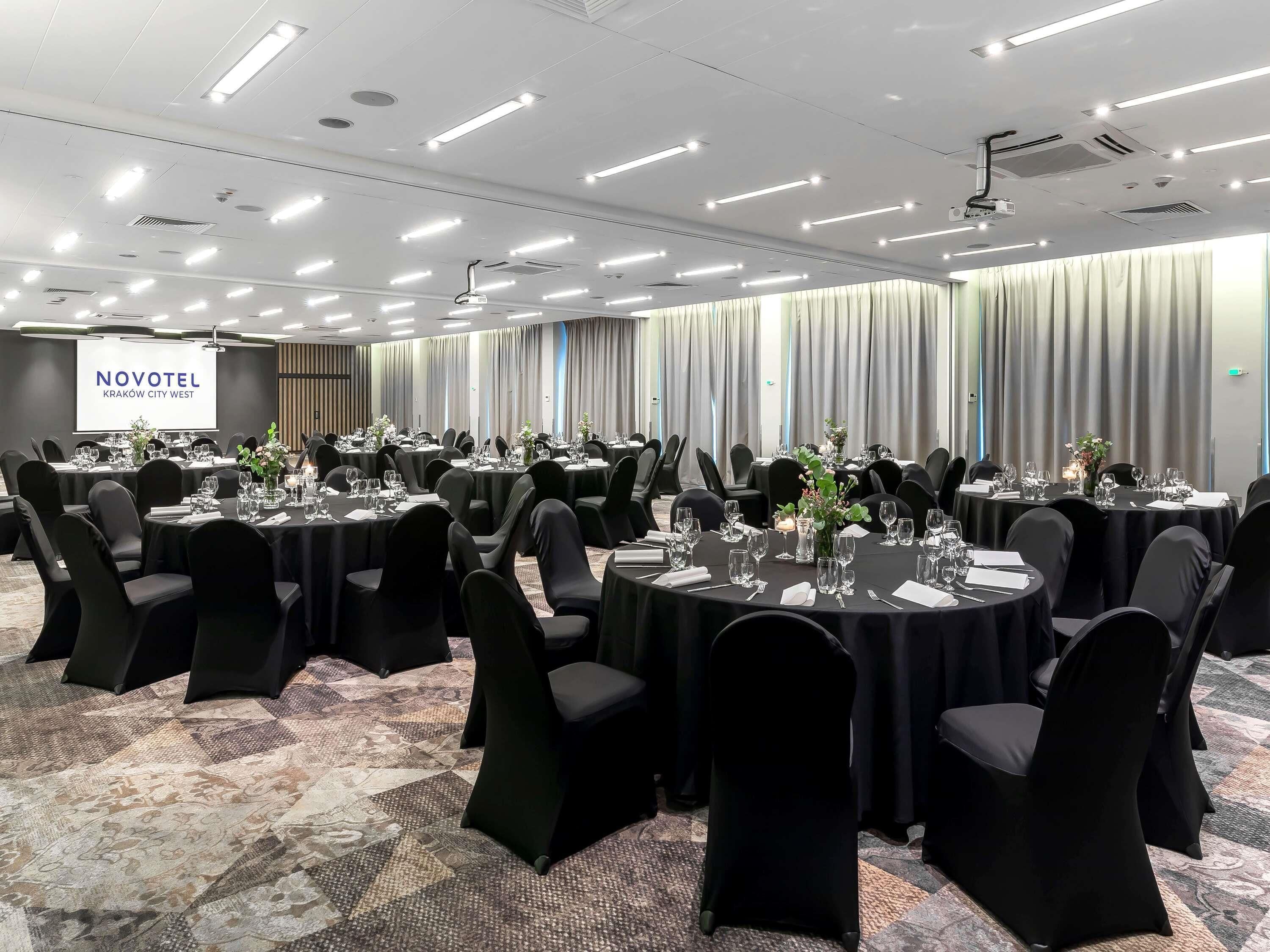 Ett konferensrum med runda bord, svarta stolar och en projektorduk som visar texten ”NOVOTEL KRAKÓW CITY WEST”.