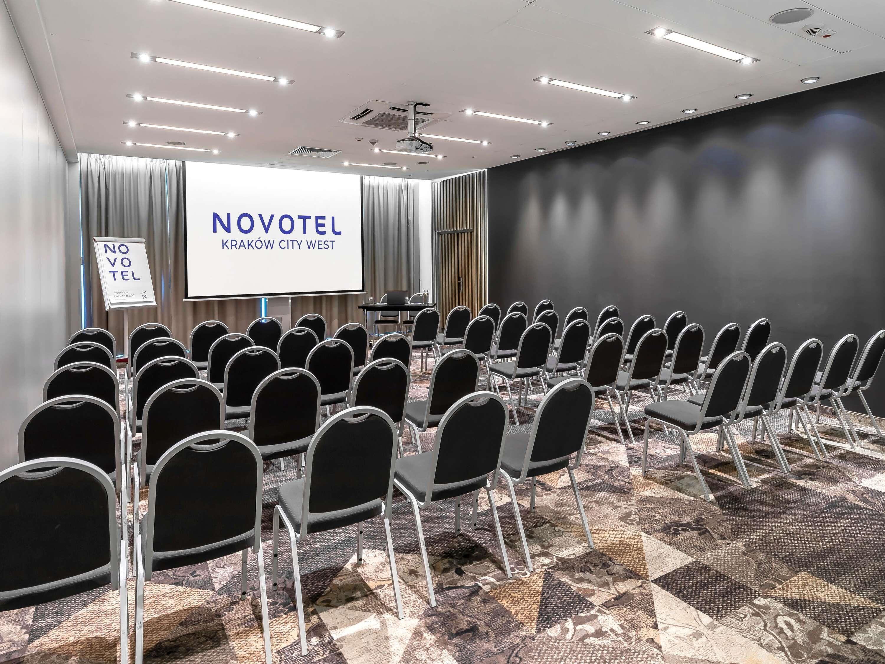 Un salón de conferencias con hileras de sillas, una pantalla de proyector y un cartel que indica Novotel Kraków City West.