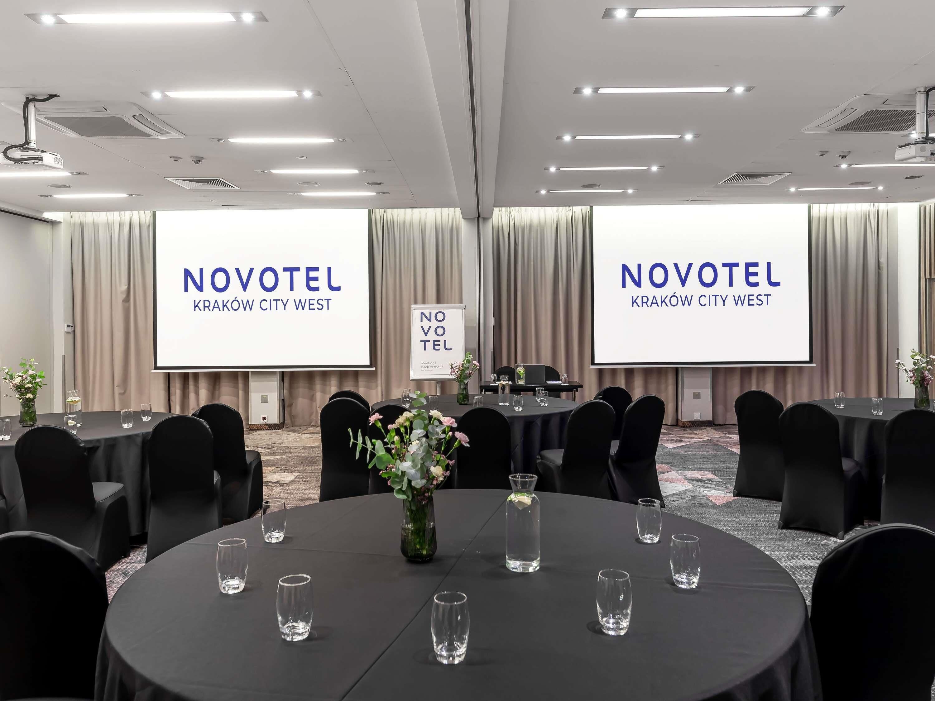 A conference room with multiple tables, chairs, and a large screen displaying 'NOVOTEL Kraków City West'.