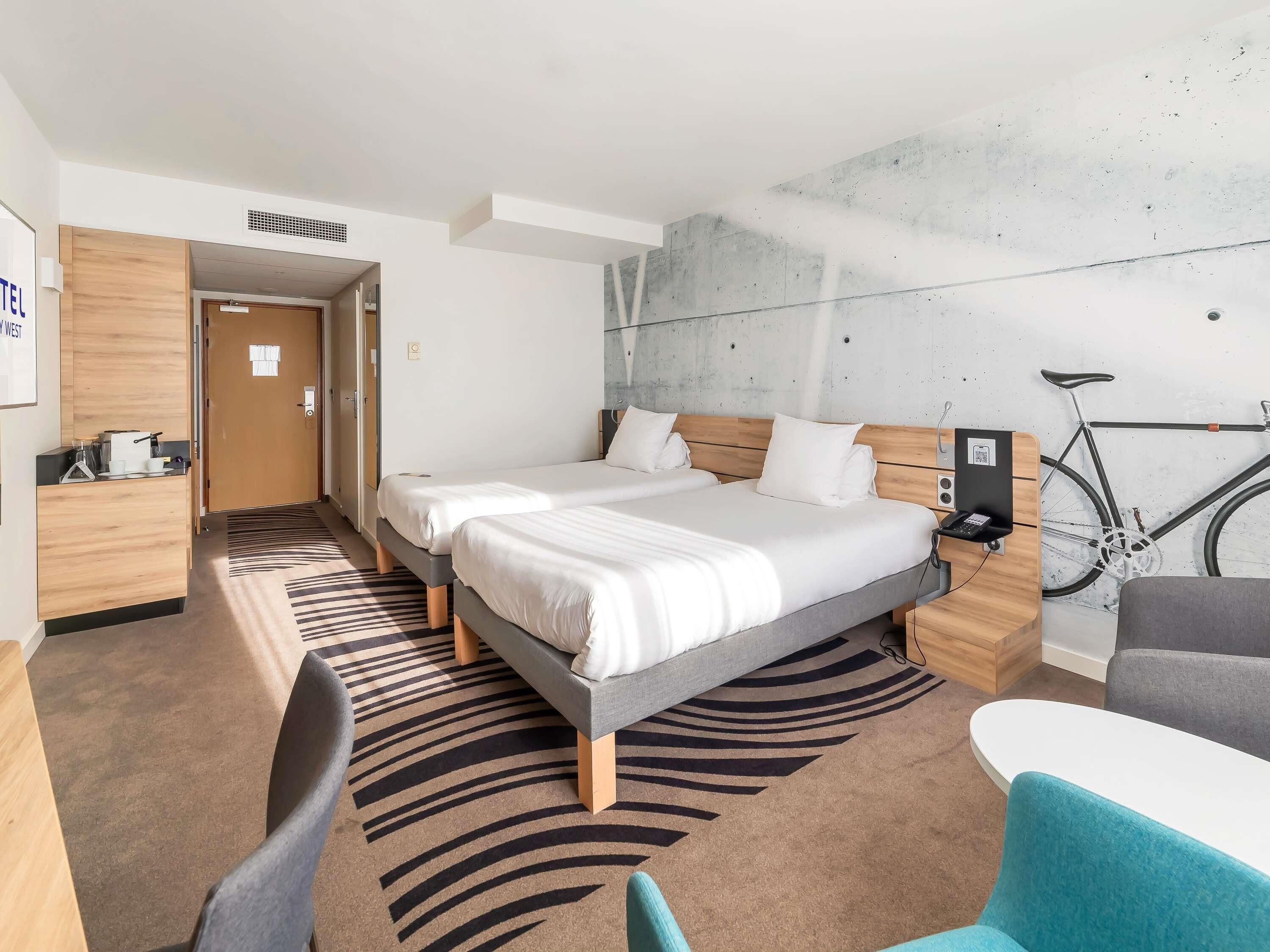 A hotel room with two beds, a desk, a chair, a bicycle, and a modern wall design.