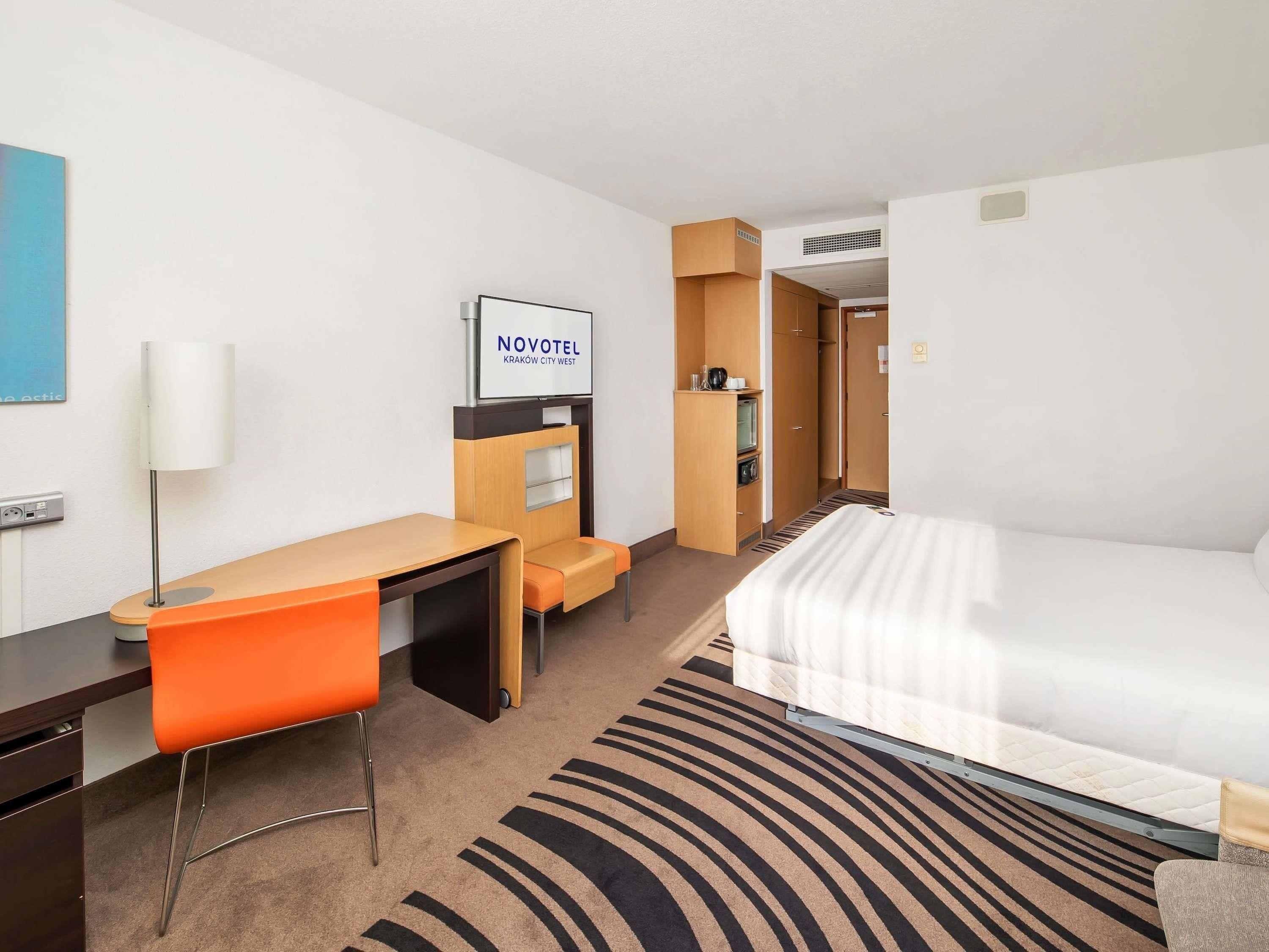Une chambre d’hôtel avec un lit, un bureau, une chaise orange, une télévision et une armoire.