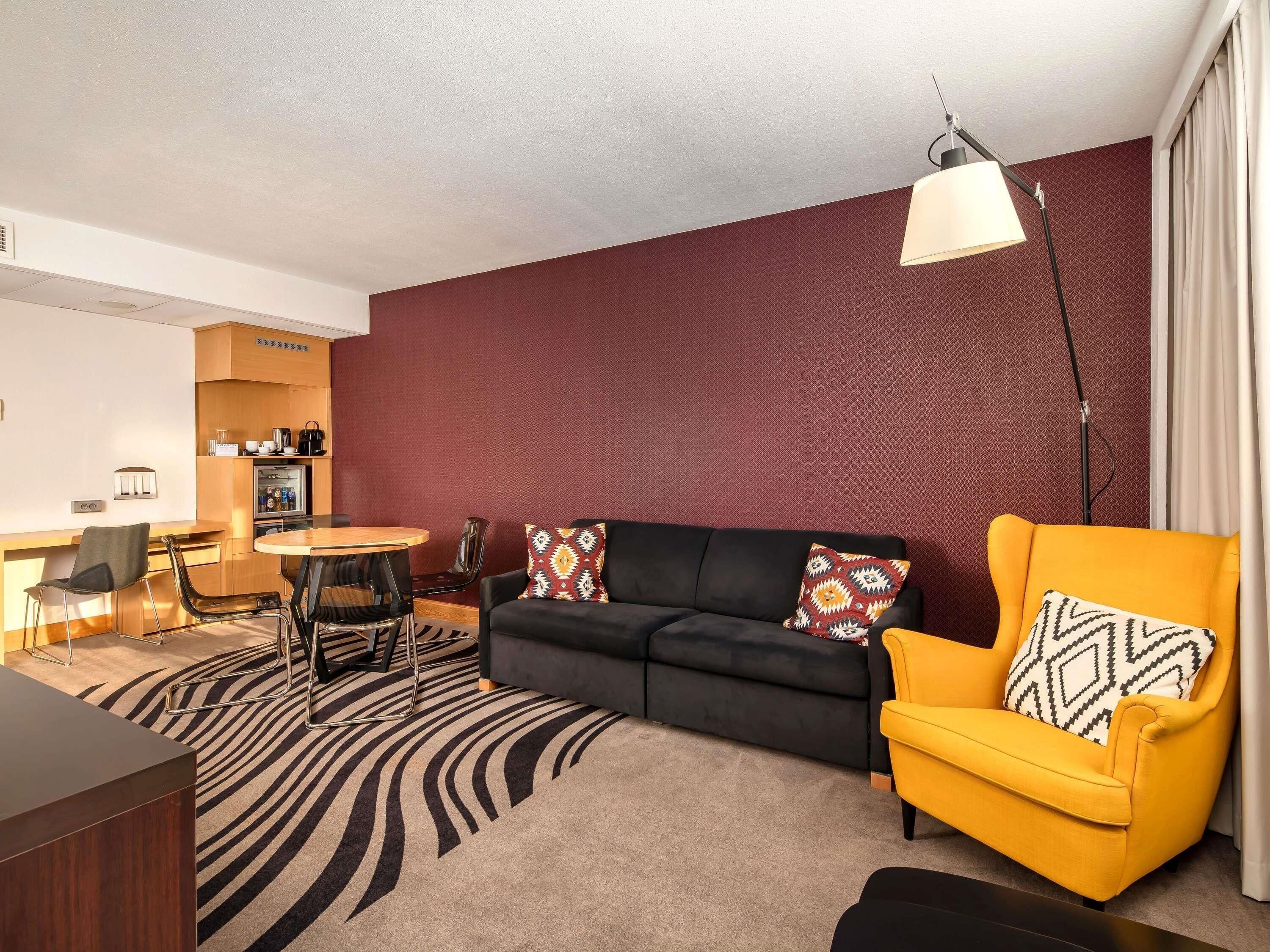 En moderne stue med en sort sofa, en gul lænestol, et rundt spisebord og en tæppe med et mønster.