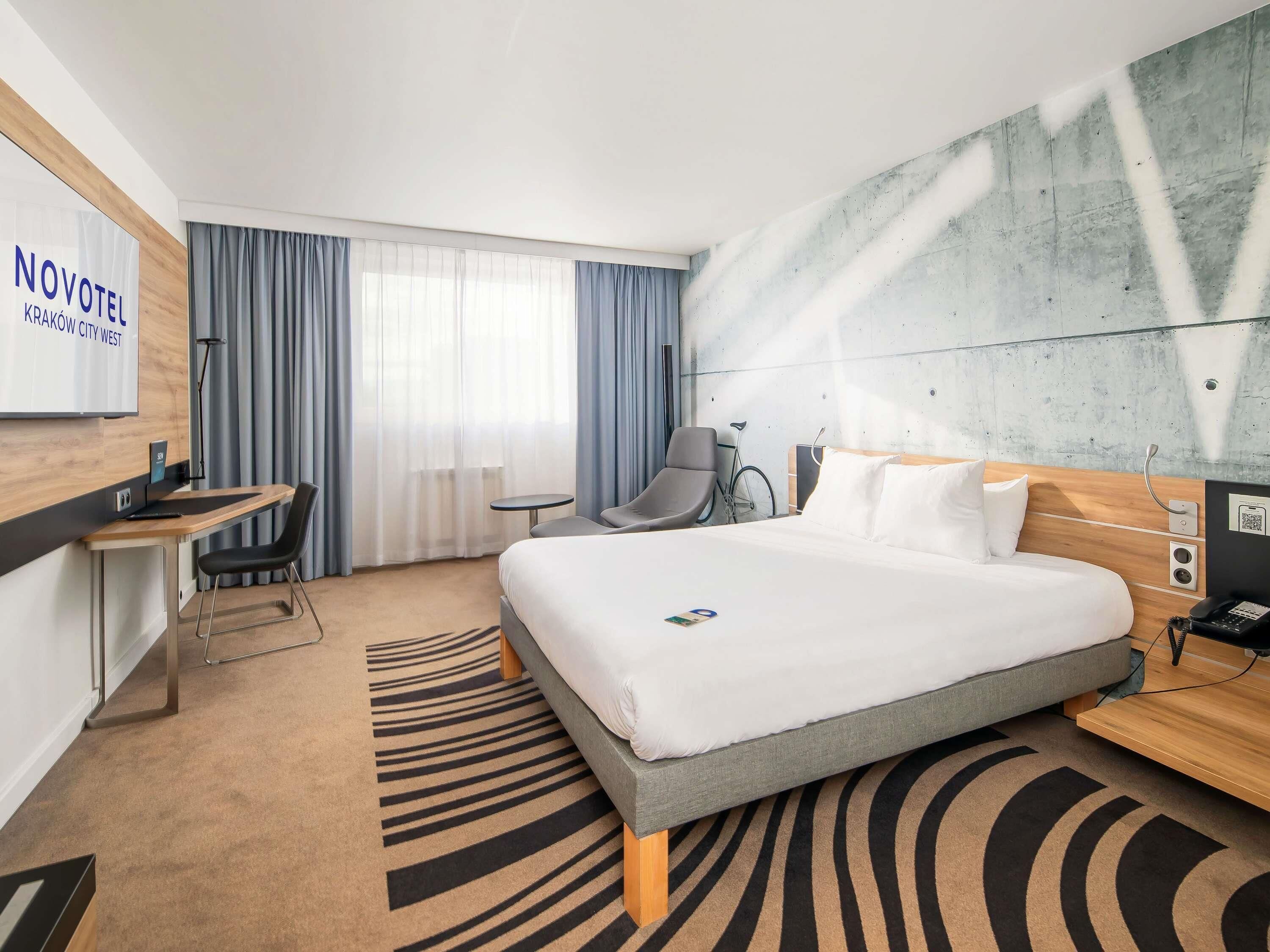 A hotel room with a bed, desk, chair, bicycle, and a large window with curtains.