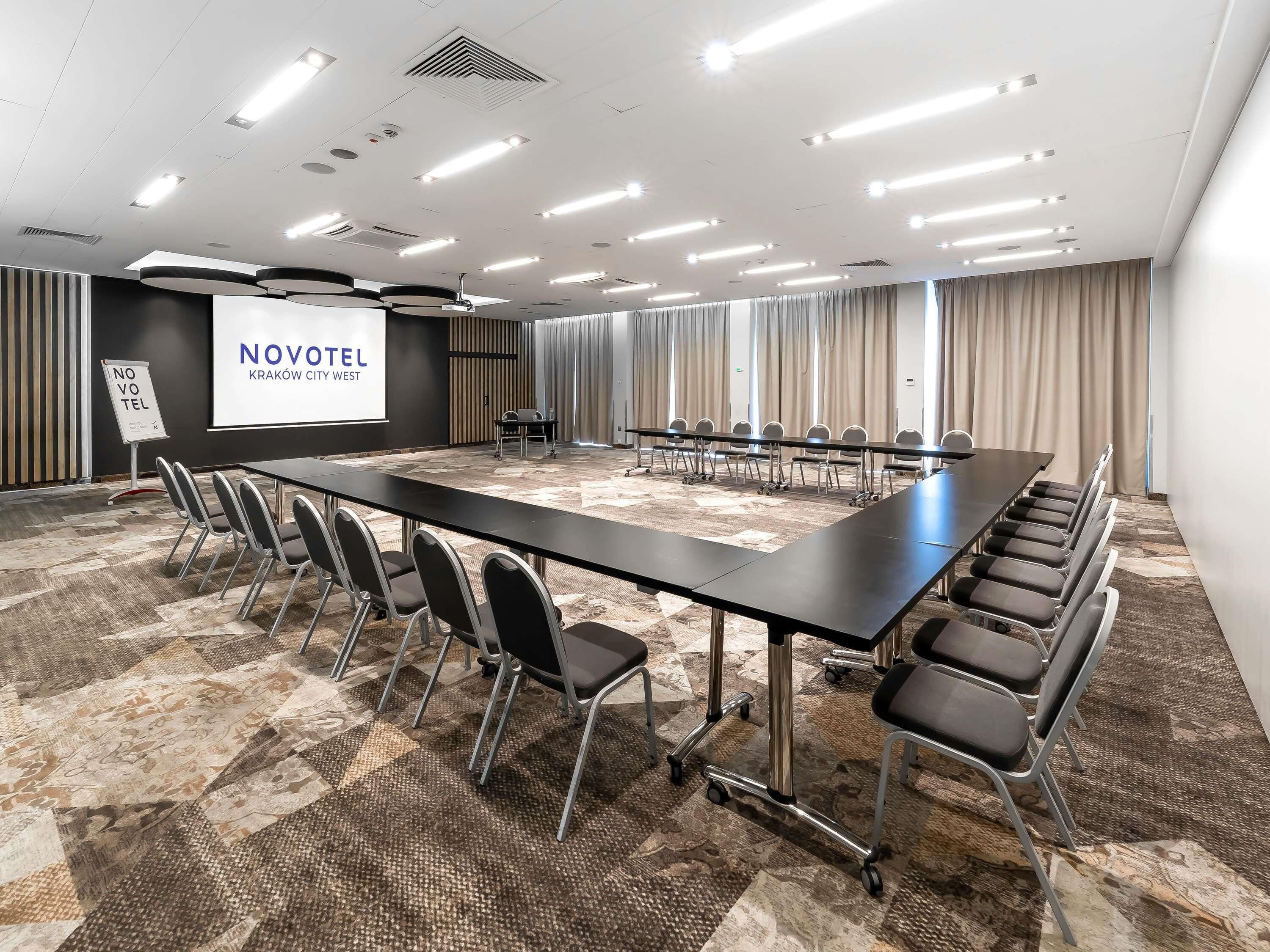 A conference room with a long table, chairs, a projector screen, and a sign for NOVOTEL Kraków City West.