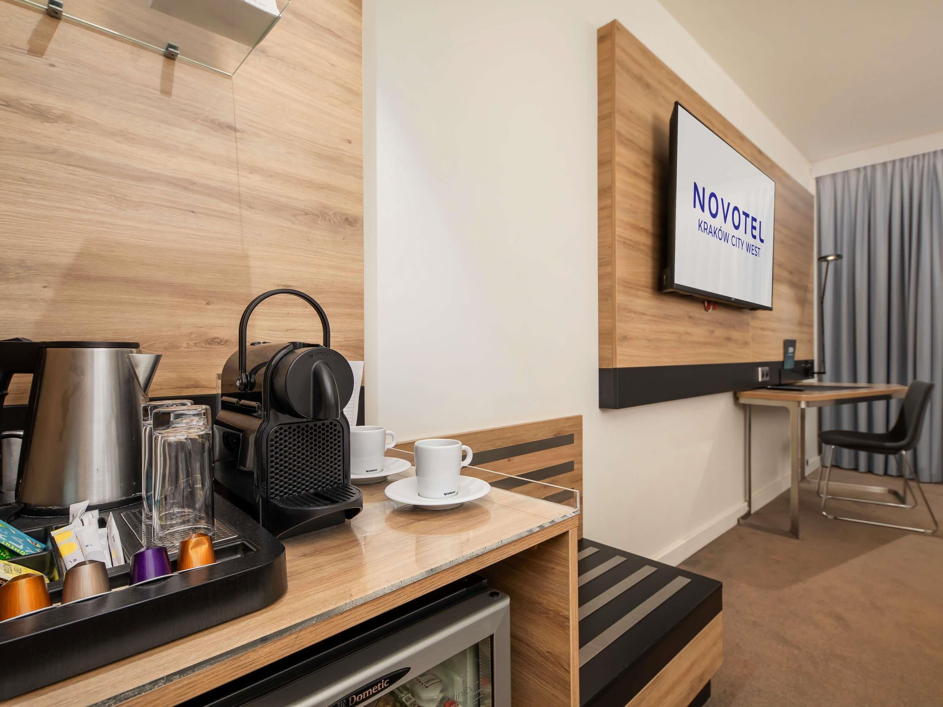 Habitación de hotel con kitchenette, cafetera, hervidor de agua, bandeja con tazas y un vaso, televisor montado en la pared y zona de estar con una silla y una mesa.