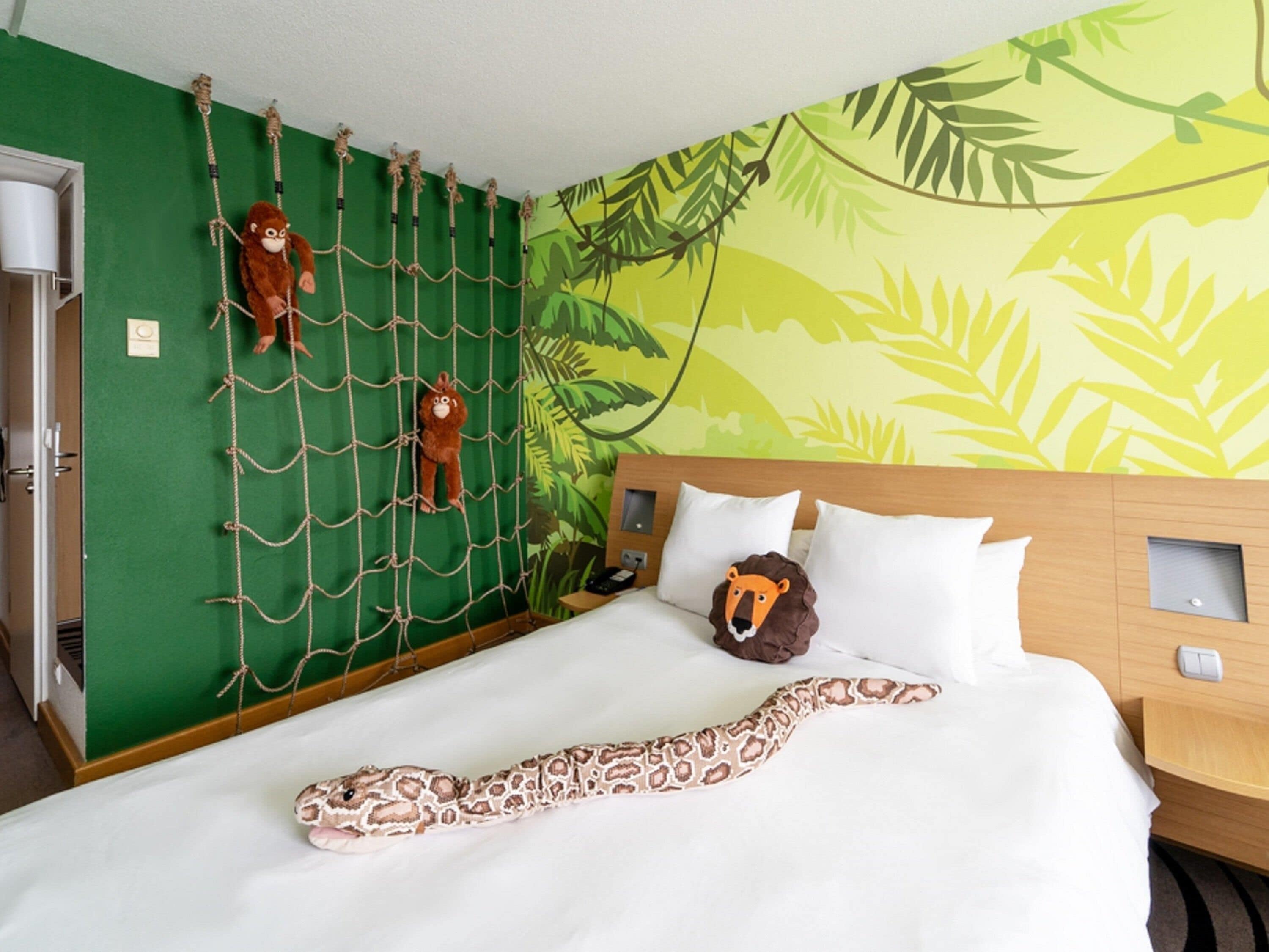 Habitación de hotel con cama, pared verde, pared de escalar con cuerdas y un mural de hojas.