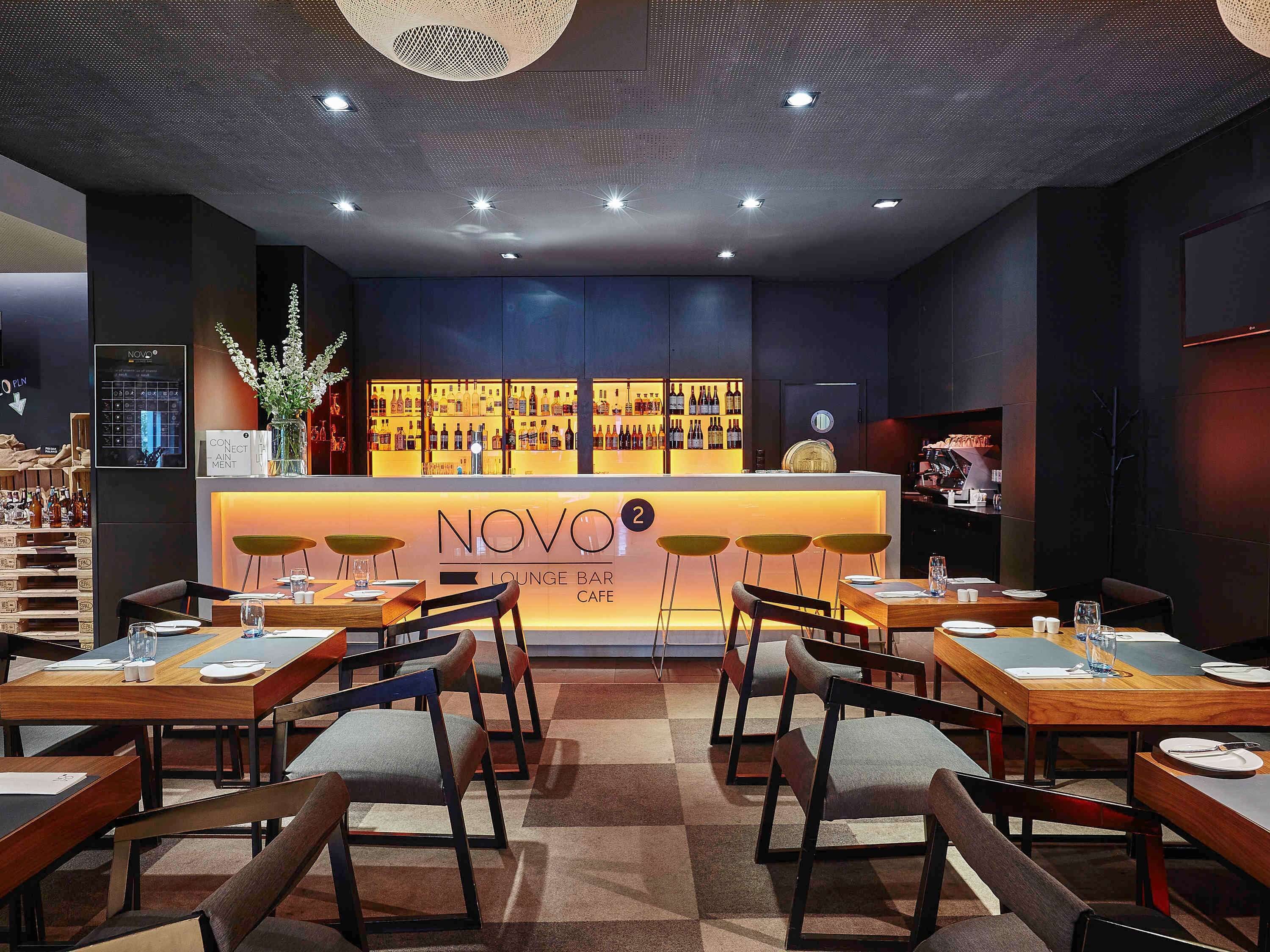 A NOVO lounge bar cafe with a bar area displaying various bottles, seating with tables and chairs, and a ceiling with recessed lighting.