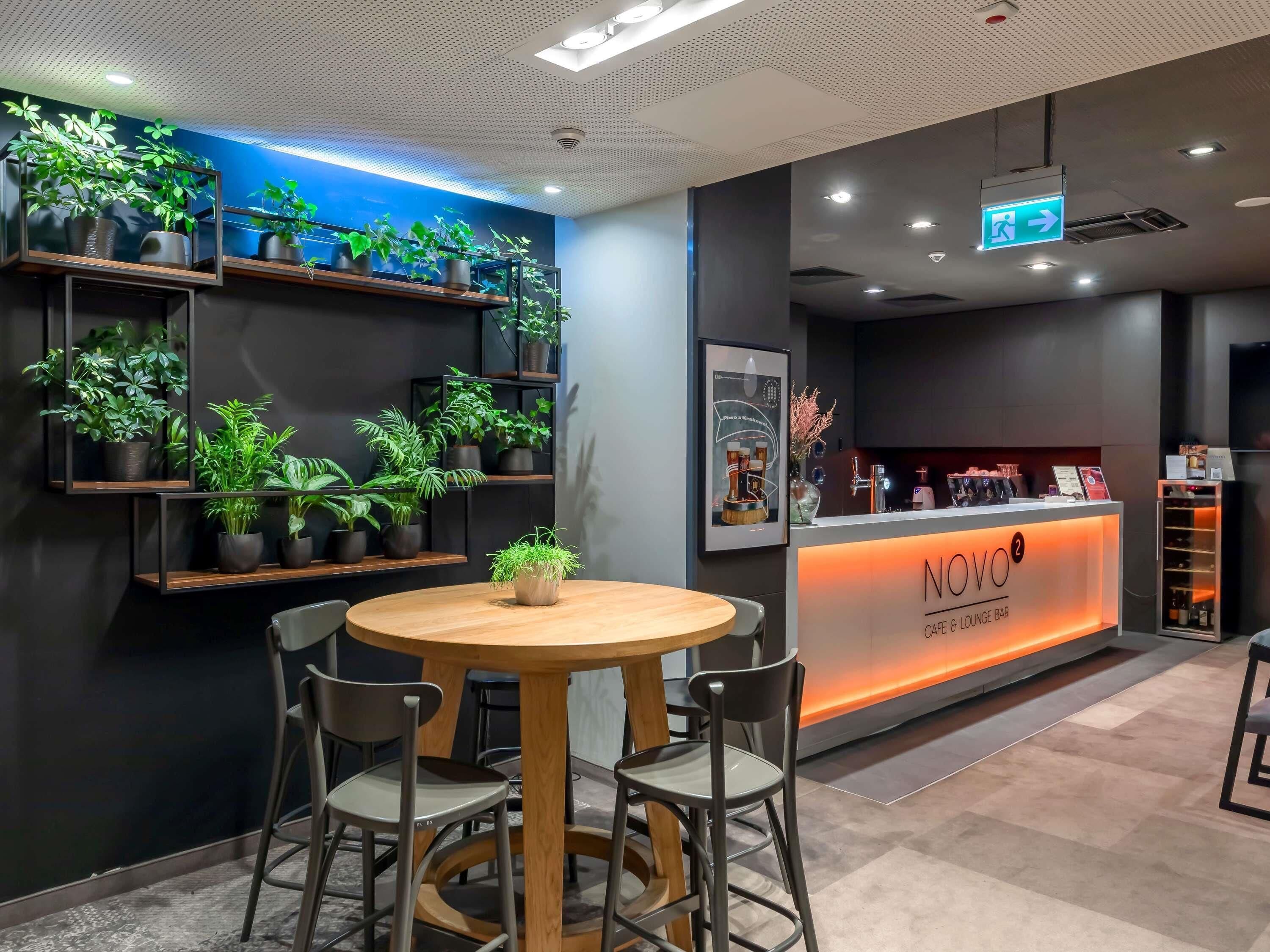 A modern cafe lounge with a round wooden table, metal chairs, and shelves with potted plants.