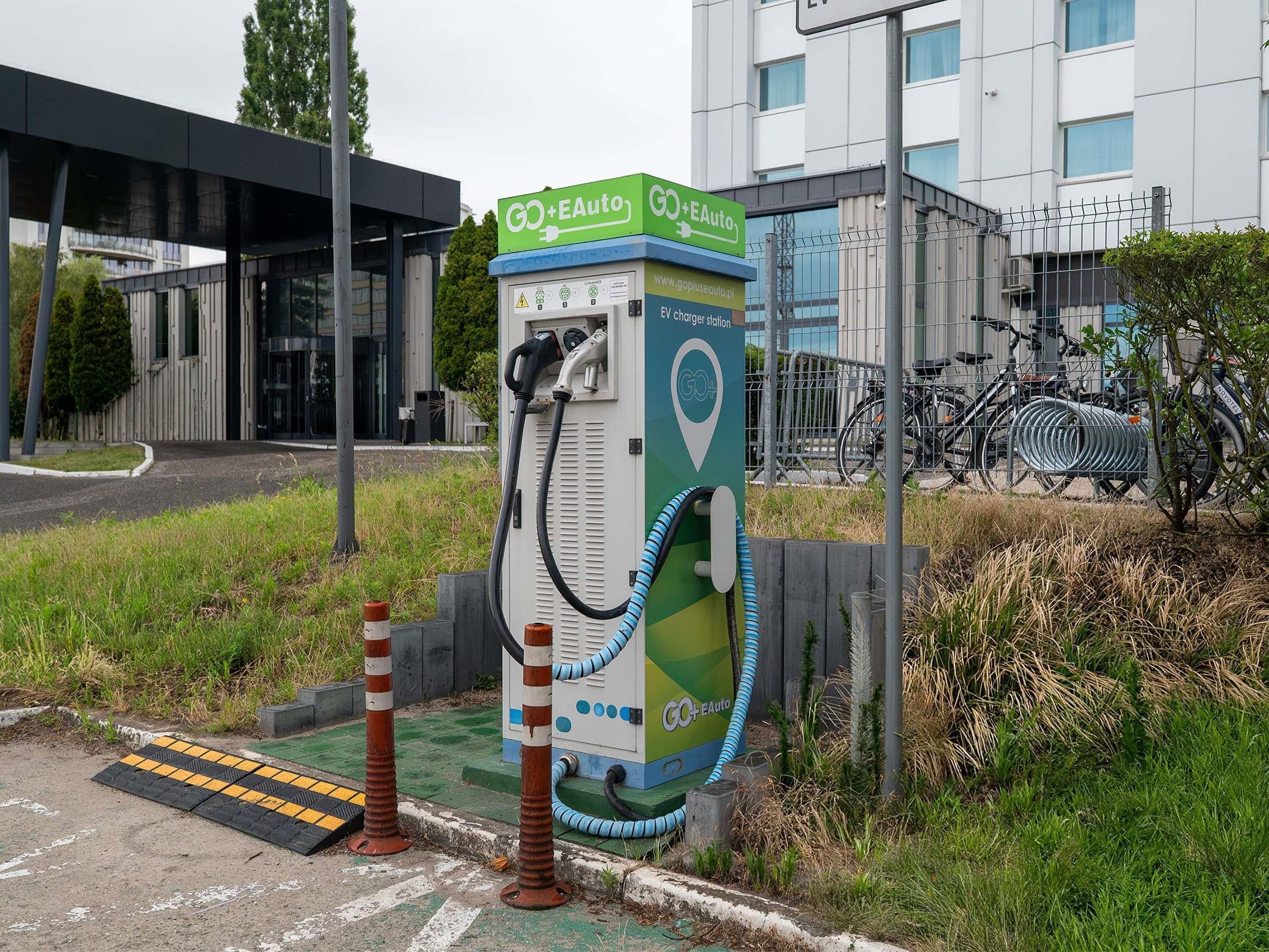 An electric vehicle charging station with a green and white color scheme, located outdoors with a building and bicycles in the background.