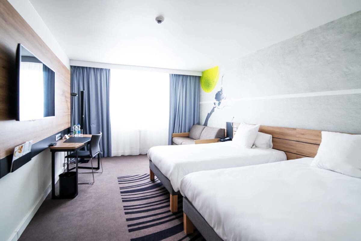 Hotelkamer met twee bedden, een bureau, een stoel, een flatscreen-tv en een groot raam met gordijnen.