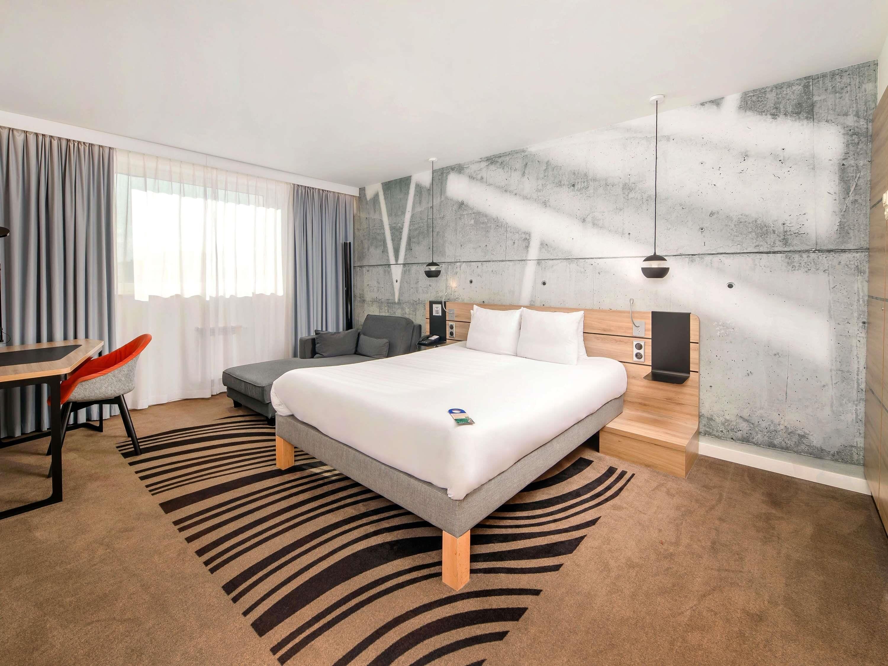 A modern hotel room with a large bed, a desk with a chair, and a sofa.