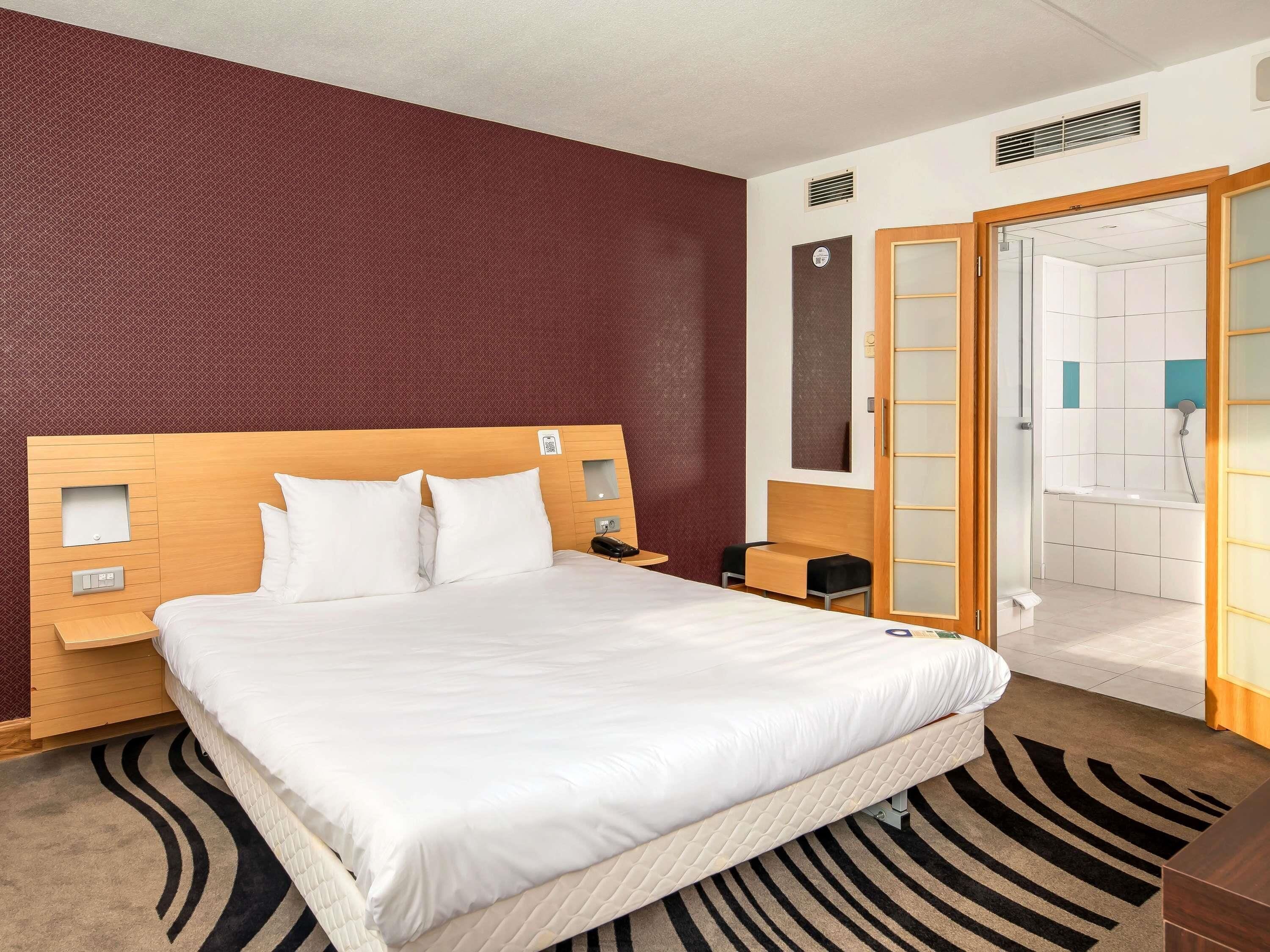 Habitación de hotel con una cama grande, cabecera de madera, mesita de noche con lámpara y baño a la vista a través de una puerta abierta.