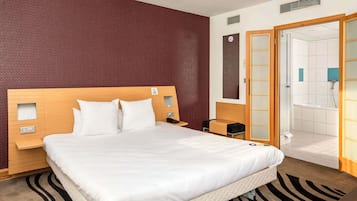 Habitación de hotel con una cama grande, cabecera de madera, mesita de noche con lámpara y baño a la vista a través de una puerta abierta.