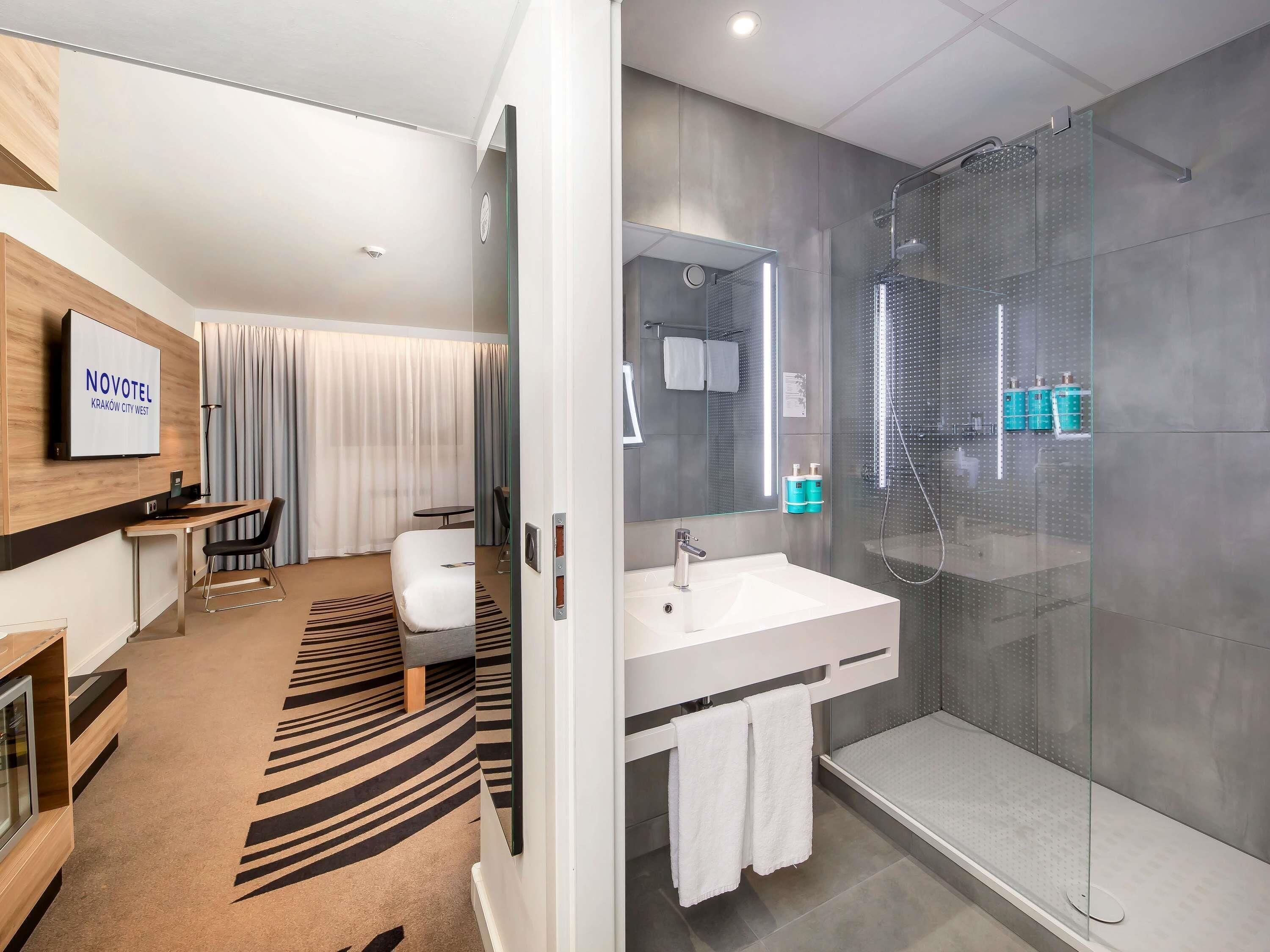 A modern hotel room with a large bed, a desk, a shower, and a TV displaying 'NOVOTEL KRAKOW CITY WEST'.