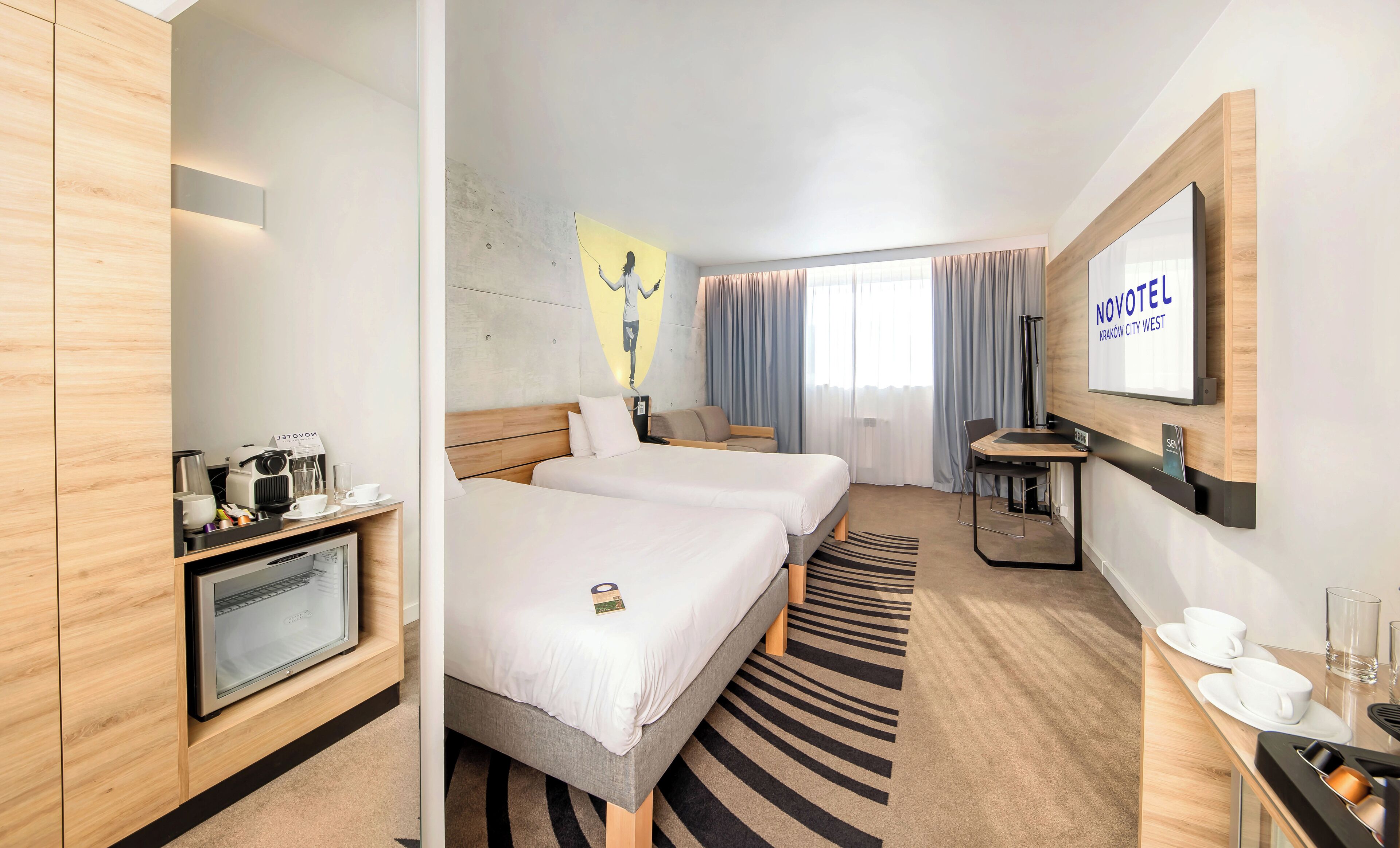 A hotel room with two beds, a desk with a coffee machine, a TV, and a wall-mounted mirror.