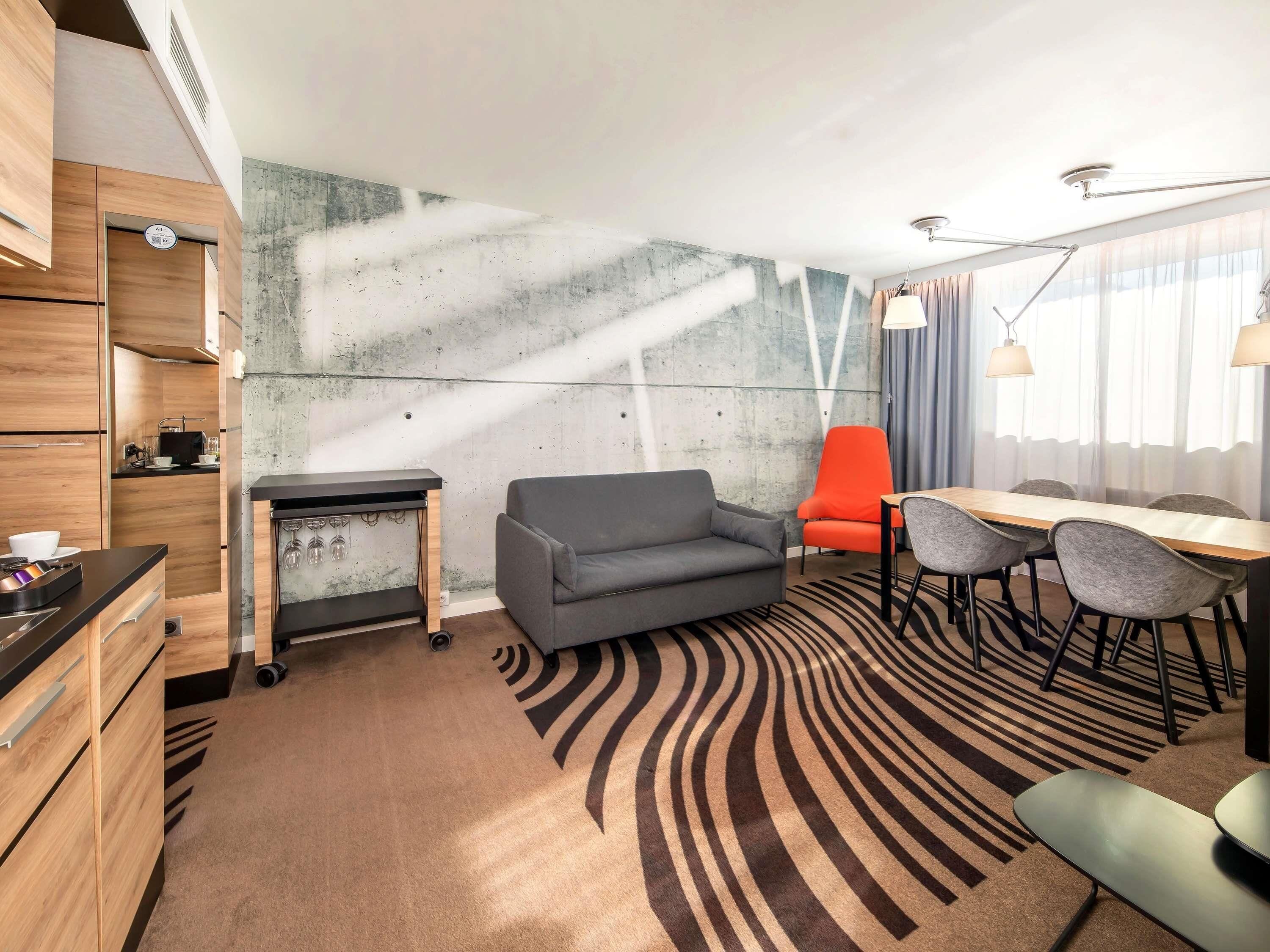Une chambre d’hôtel moderne dotée d’une kitchenette, d’un canapé gris, d’un fauteuil orange, d’une table à manger avec des chaises et d’un tapis à motifs.