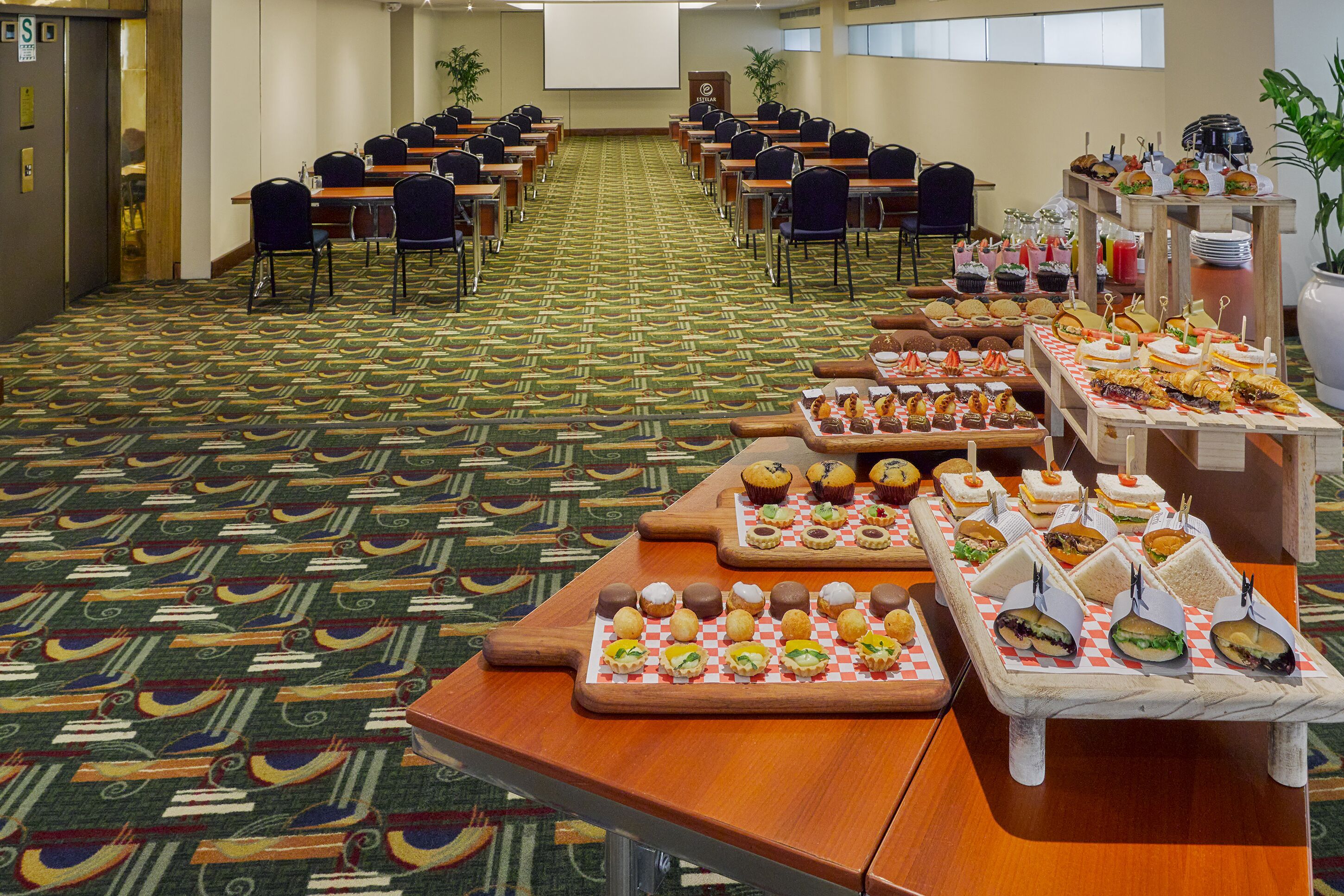 A conference room with rows of tables and chairs, a buffet table with various food items, and a projector screen on the wall.
