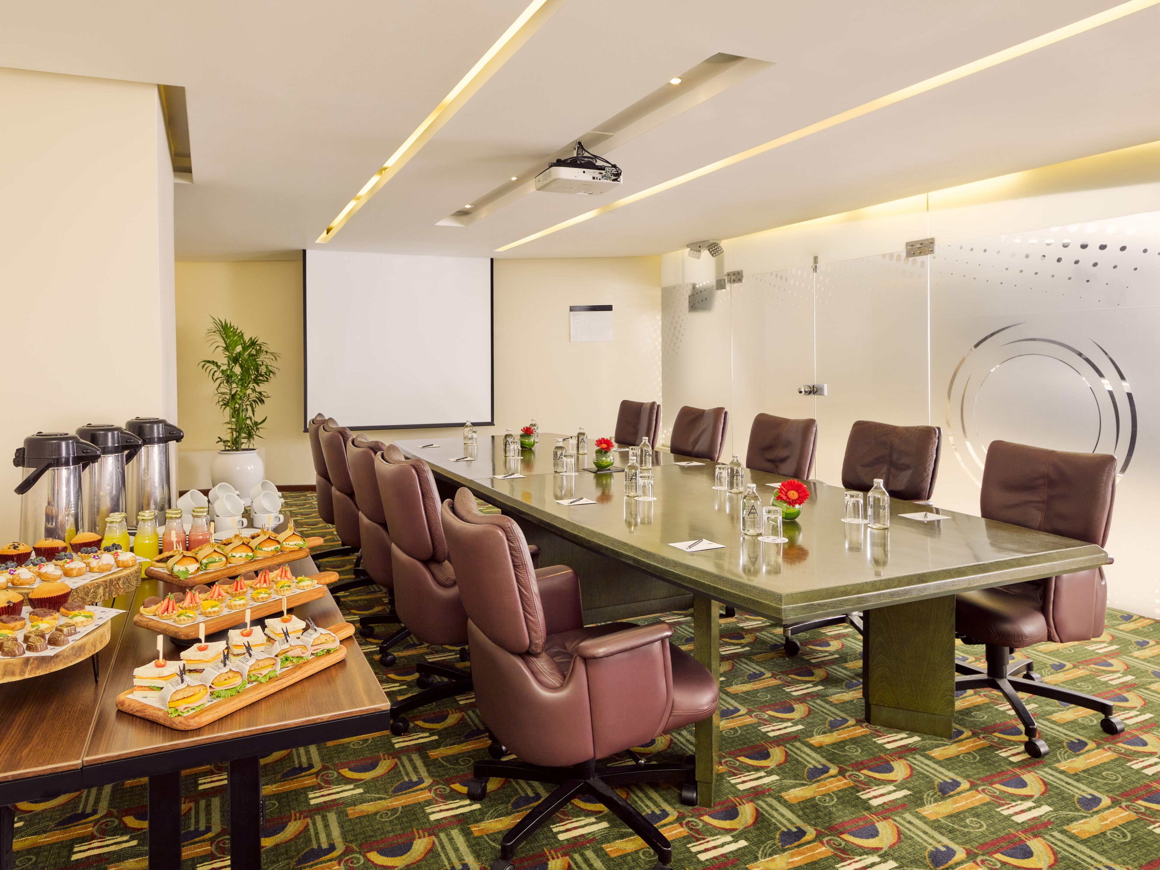 A conference room with a long table, brown leather chairs, a projector screen, and a buffet with various snacks.