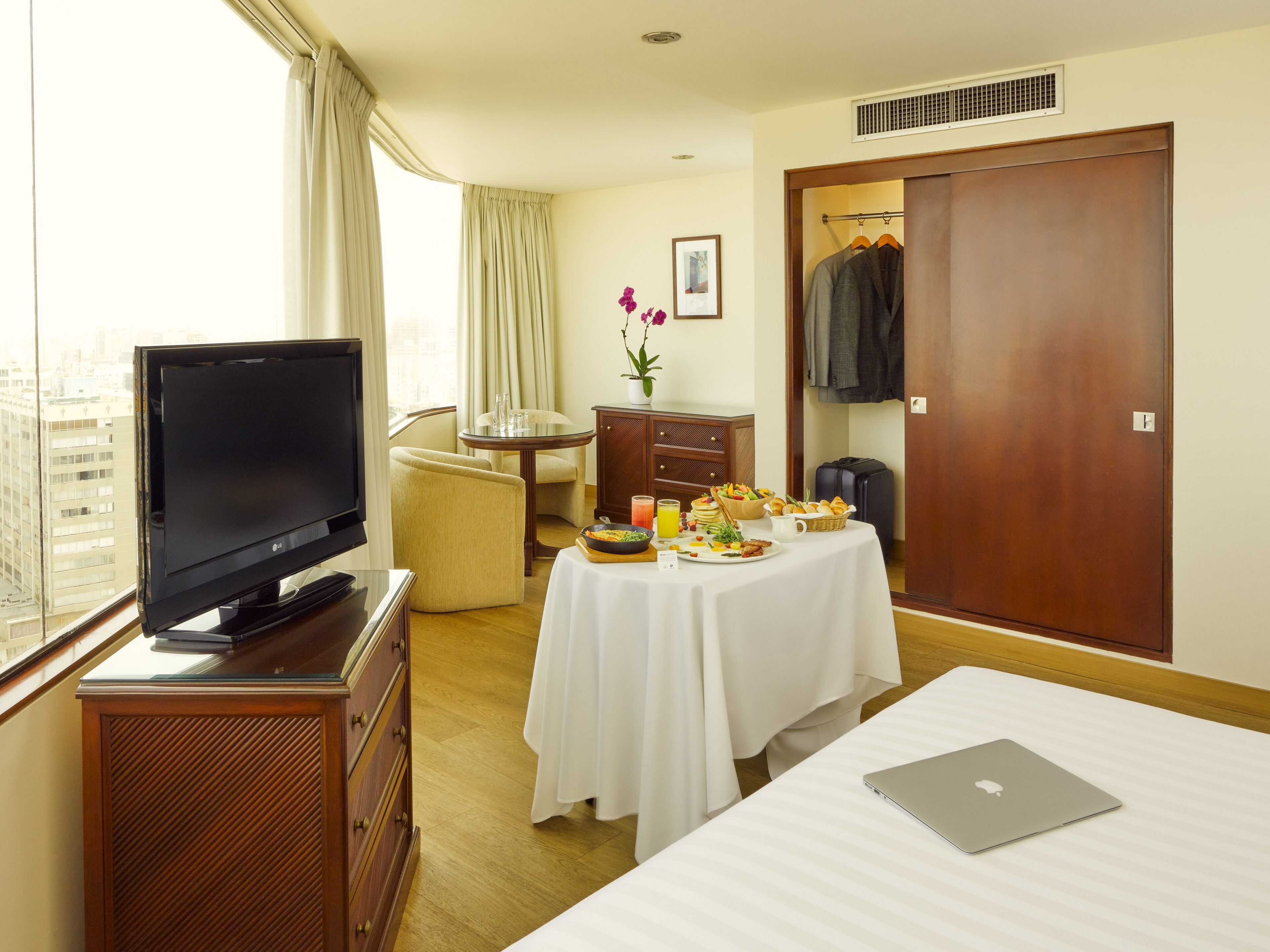 A hotel room with a bed, a desk with a laptop, a TV on a wooden cabinet, a dining table with breakfast, a wardrobe, and a window with curtains.