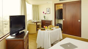 A hotel room with a bed, a desk with a laptop, a TV on a wooden cabinet, a dining table with breakfast, a wardrobe, and a window with curtains.