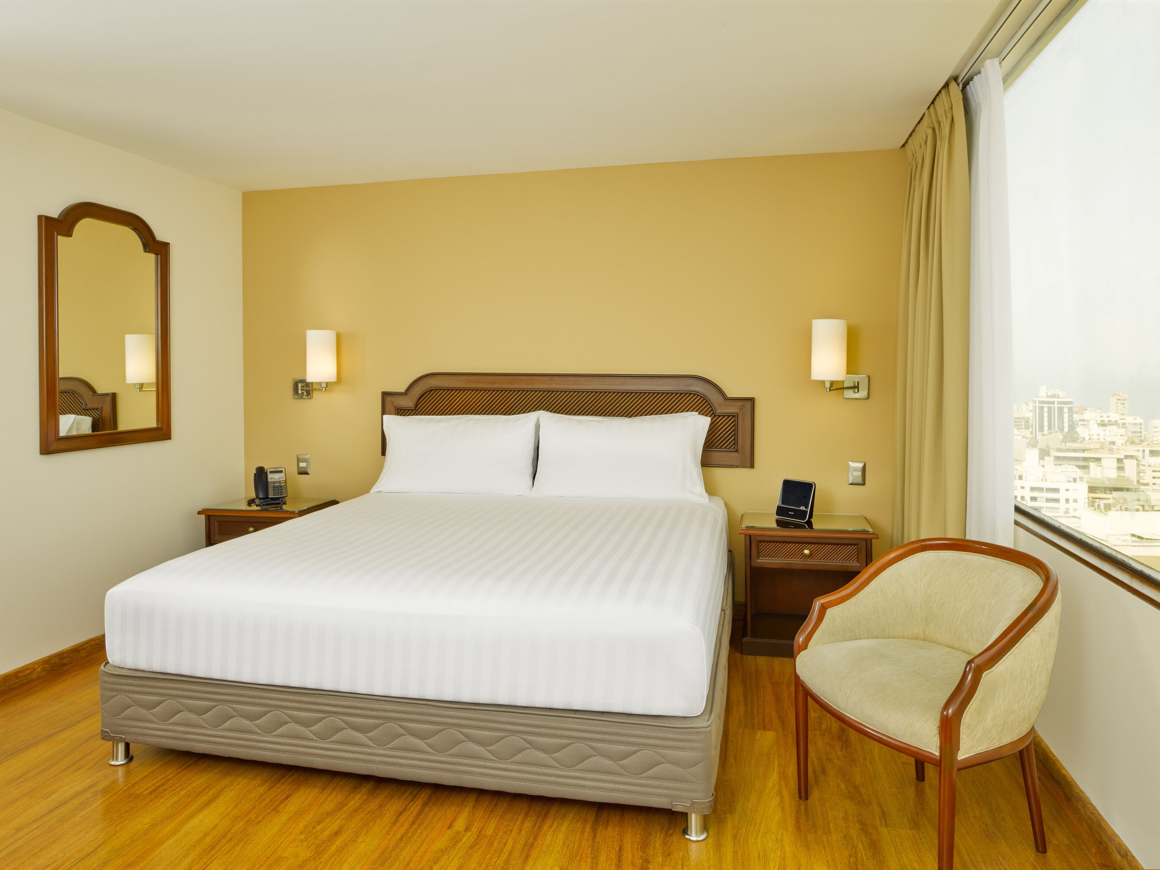 A hotel room with a large bed, bedside tables, a chair, a mirror, and a view of the city through the window.