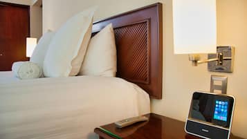 Quarto de hotel com cama, almofadas, controle remoto e estação de carregamento para smartphone.
