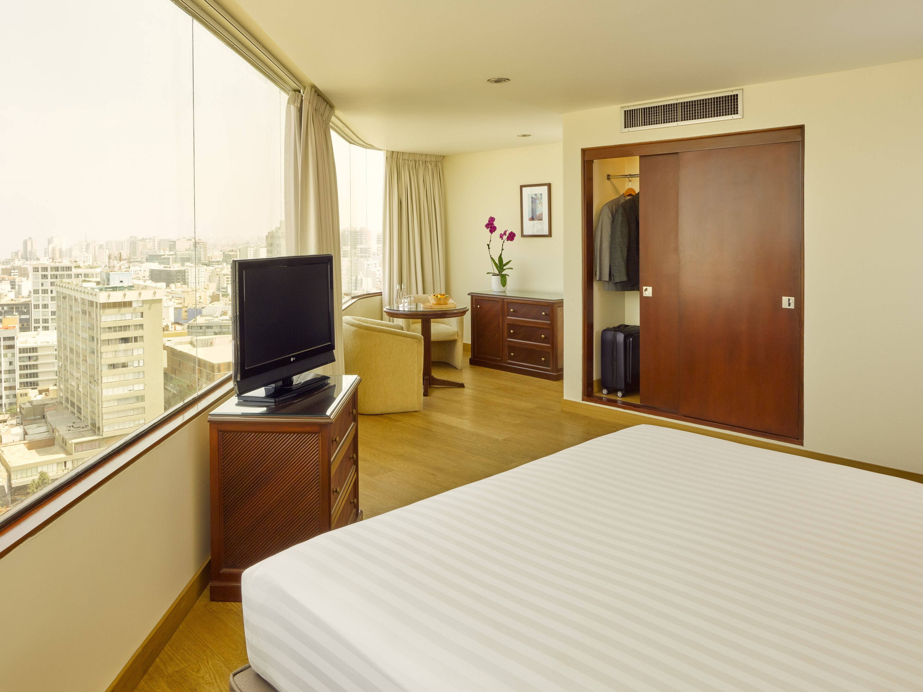 A hotel room with a bed, a TV on a wooden stand, a desk with a chair, a wardrobe, and a view of the city through large windows.