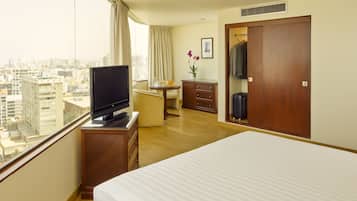 A hotel room with a bed, a TV on a wooden stand, a desk with a chair, a wardrobe, and a view of the city through large windows.