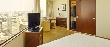 Una habitación de hotel con una cama, un televisor sobre un soporte de madera, un escritorio con una silla, un armario y vistas de la ciudad a través de amplias ventanas.