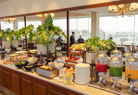 A buffet with various food items and drink dispensers, surrounded by potted plants and large windows offering a city view.