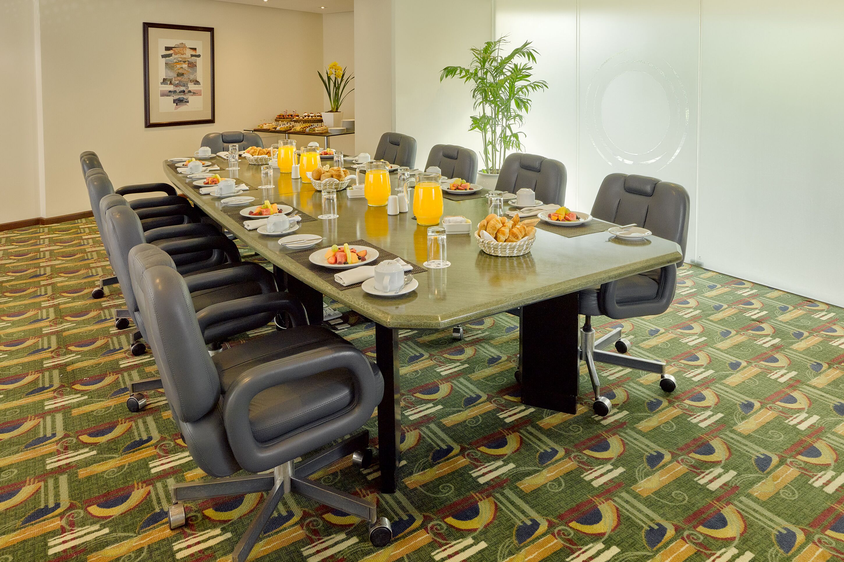 A conference room with a long table set for breakfast, including plates, cups, and juice. There are chairs around the table, and a plant is visible in the background.