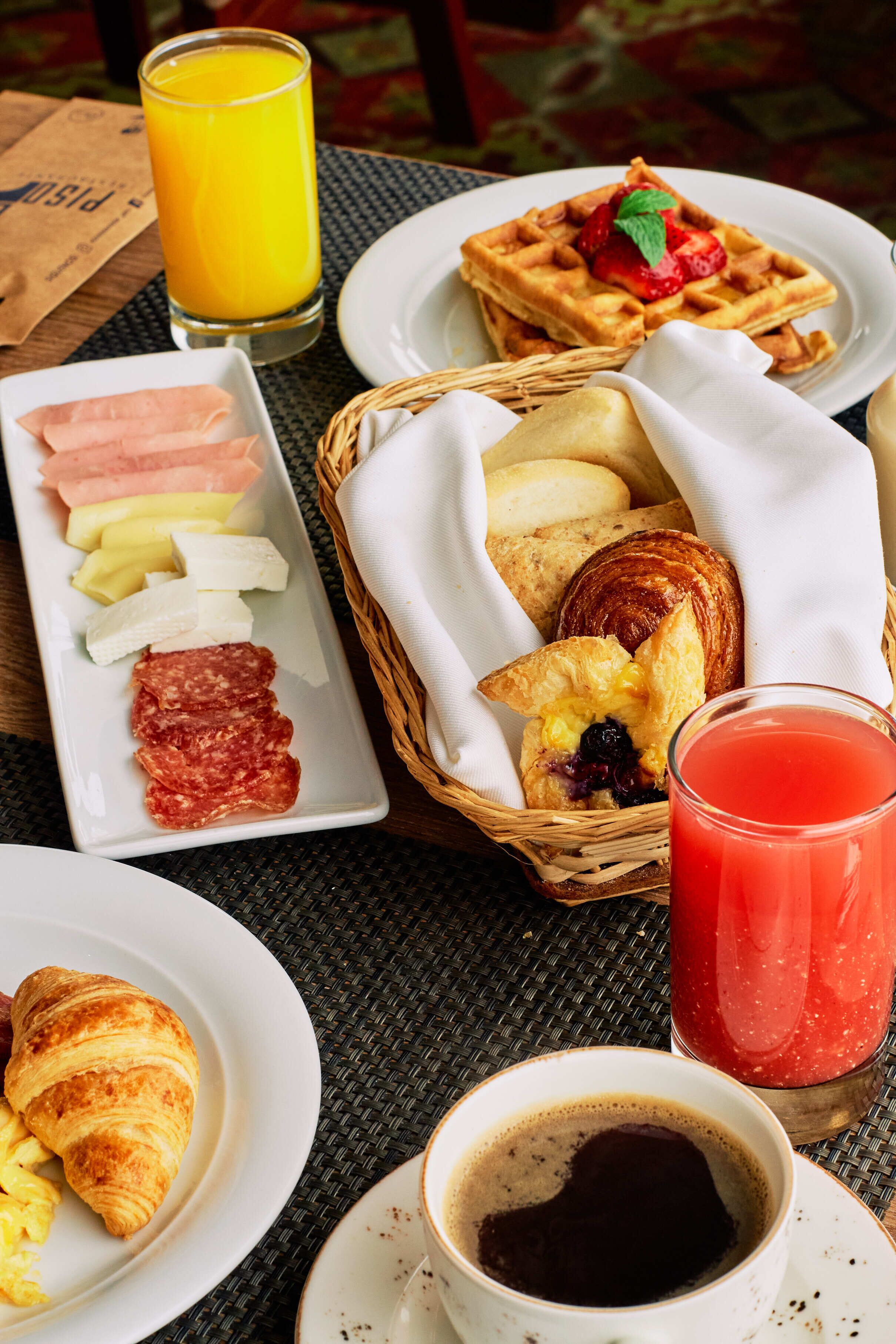 A breakfast setting with a plate of ham, cheese, and fruit, a basket of pastries, a waffle with a strawberry, and a glass of orange juice.