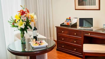 A hotel room with a round glass table, a wooden desk, a chair, a bed, a framed picture on the wall, and a window with white curtains.
