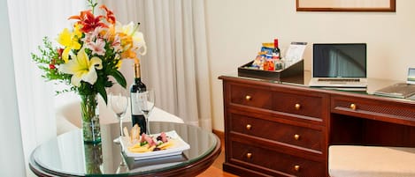 Habitación de hotel con una mesa redonda de cristal, un escritorio de madera, una silla, una cama, un cuadro en la pared y una ventana con cortinas blancas.