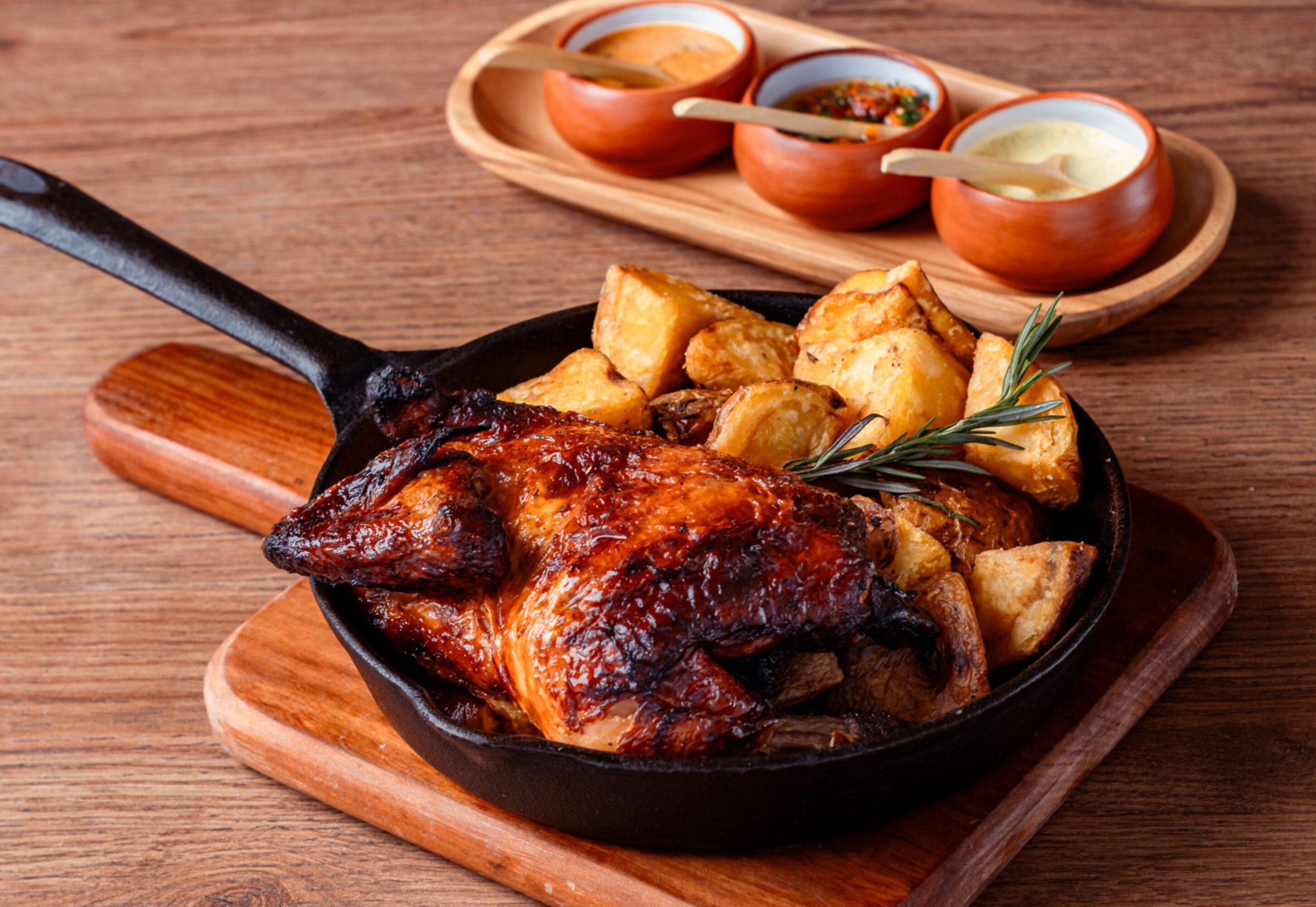 Pollo asado en una sartén de hierro fundido, junto a patatas asadas y romero, acompañado de dos recipientes pequeños con salsas sobre una tabla de madera.