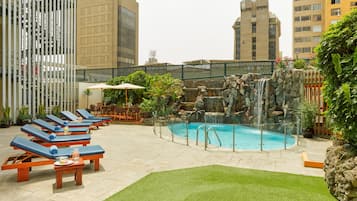A rooftop pool area with lounge chairs, a small waterfall feature, and a view of surrounding buildings.