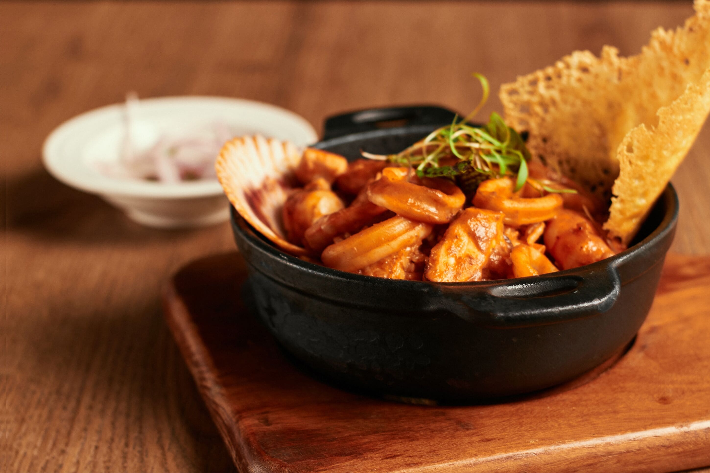 A black bowl of food with a side of sliced onions and a piece of bread on a wooden table.