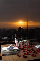 Una mesa dispuesta para dos, con una botella de vino, una servilleta y un plato de vidrio. La palabra “LOVE” está escrita con letras, y hay pétalos de rosa esparcidos sobre la mesa.