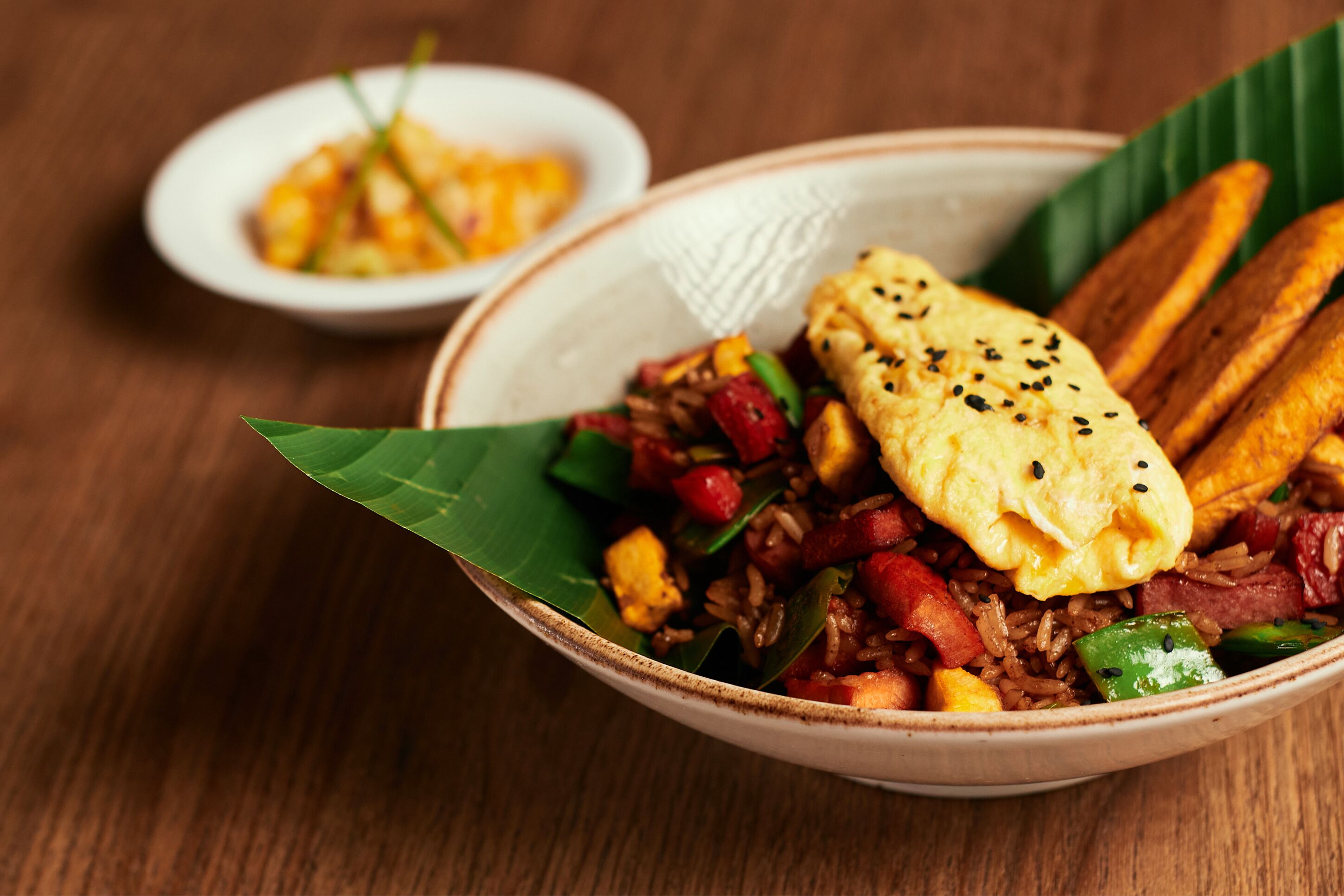 A bowl of fried rice with vegetables and a fried egg on a banana leaf, accompanied by a small bowl of soup and fried plantains.