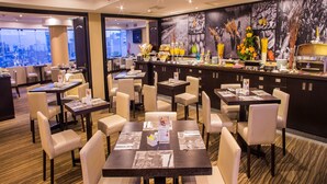 Un comedor de hotel con varias mesas y sillas, una barra de buffet y vistas a la ciudad a través de amplias ventanas.